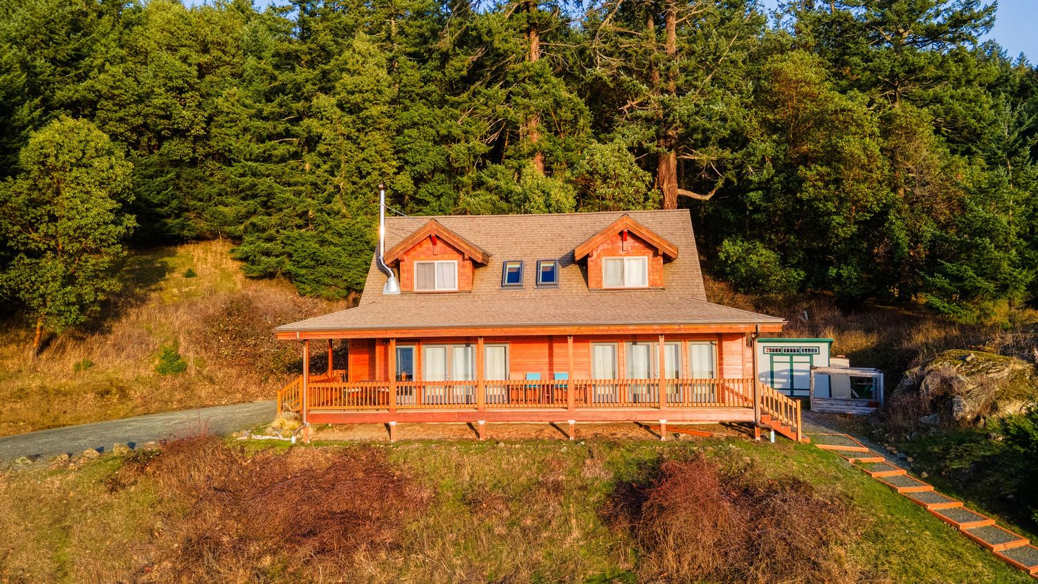 Red waterfront house with wrap-around deck nestled among evergreen trees on hillside.