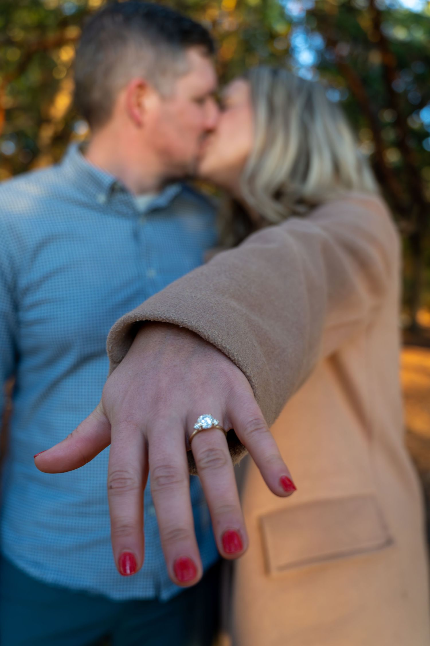 Close-up of an engagement ring on a hand with red nail polish while couple embraces in background.