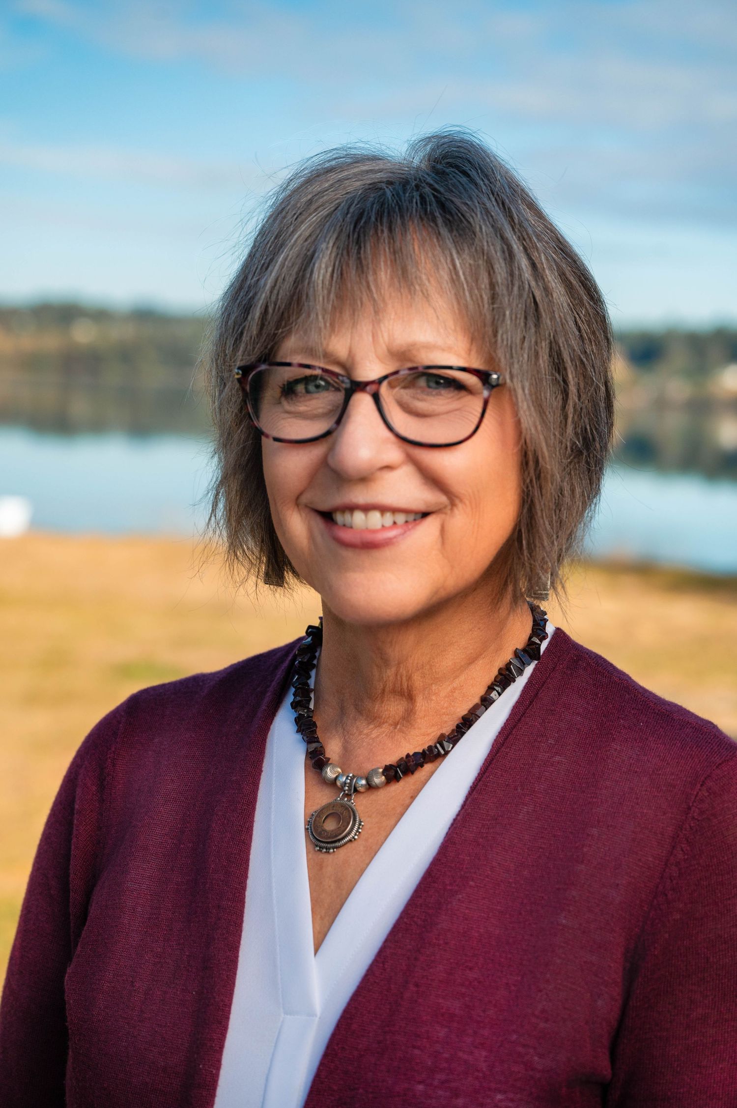 Outdoor portrait of person in burgundy cardigan and glasses with water and nature in background.
