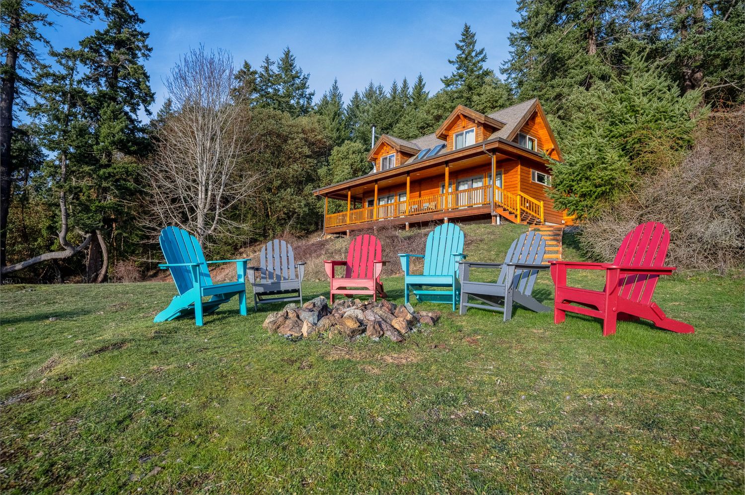 Colorful Adirondack chairs arranged around a fire pit in front of a luxury log cabin.