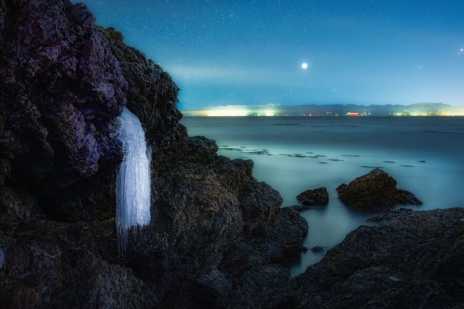 Moonlit waterfall cascades down rocky cliffs into a misty ocean cove under a starry night sky.