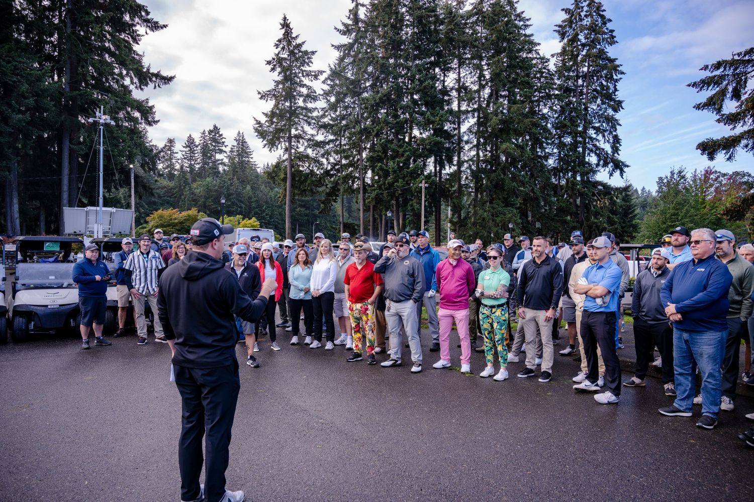 Large crowd gathered in parking lot surrounded by tall pine trees.