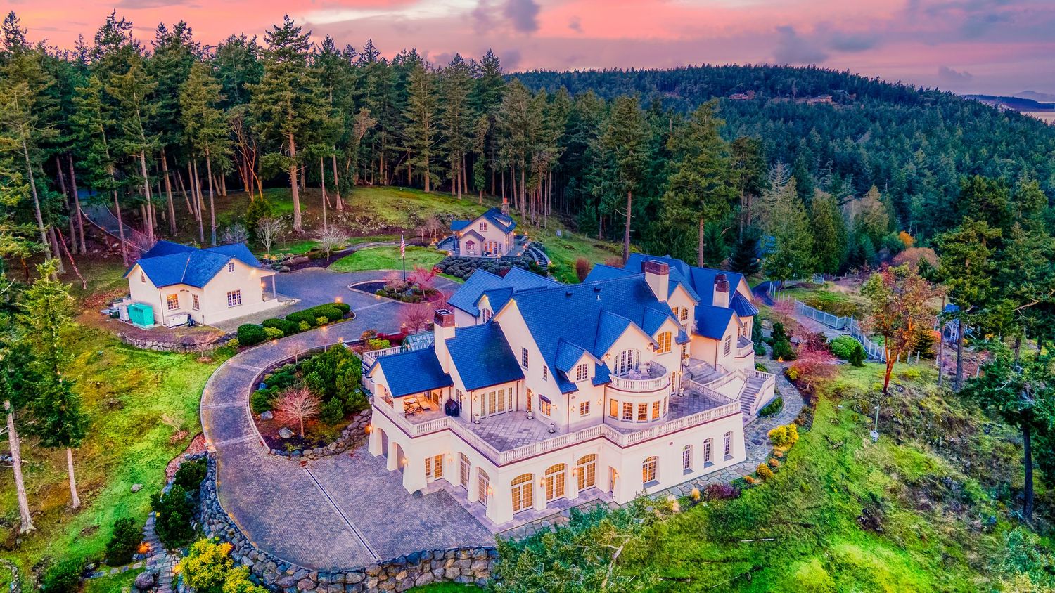 Aerial view of grand Victorian-style mansion with blue roofs surrounded by evergreen forest at sunset.