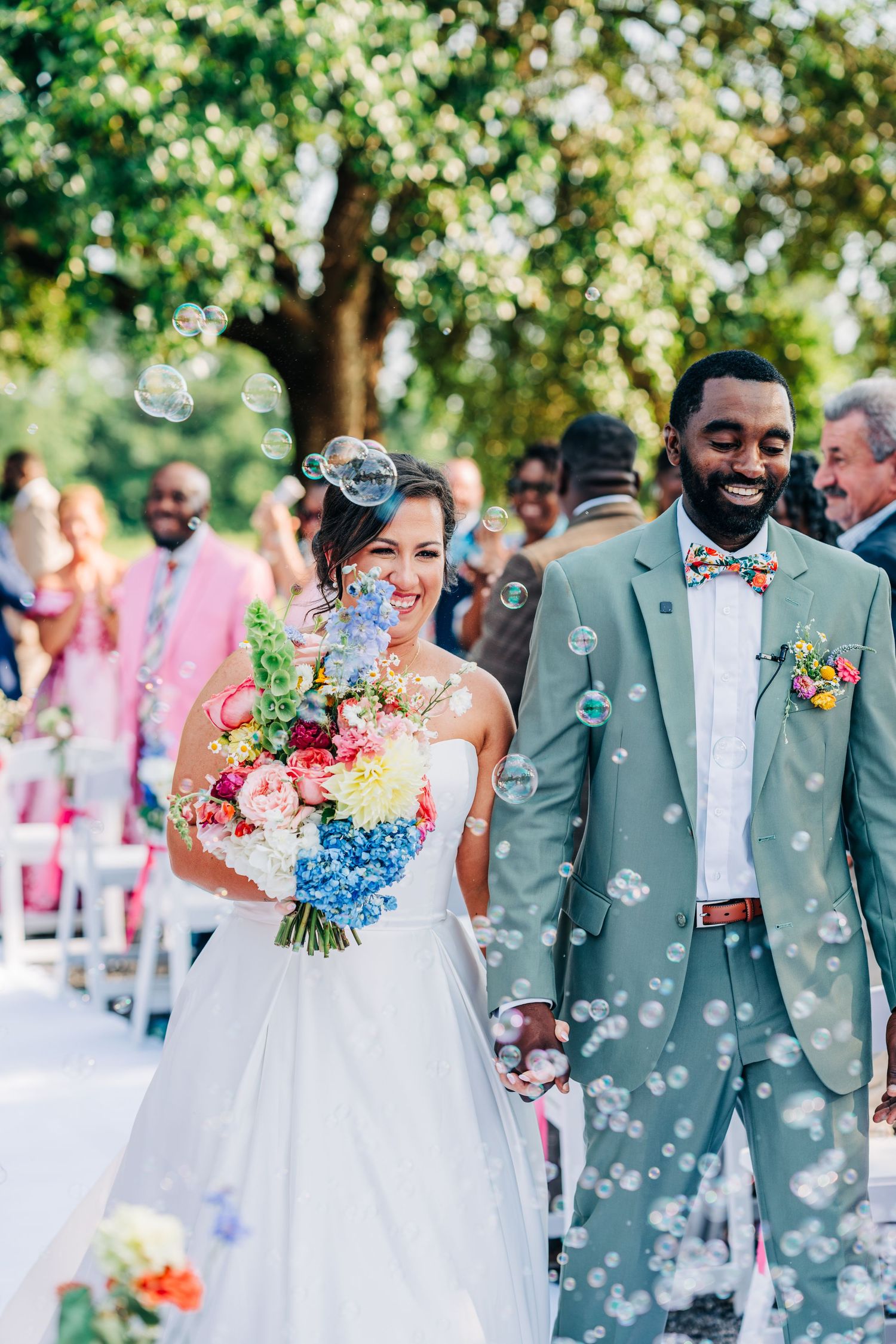 Wedding party celebration with bubbles floating through air as couple walks down outdoor aisle.