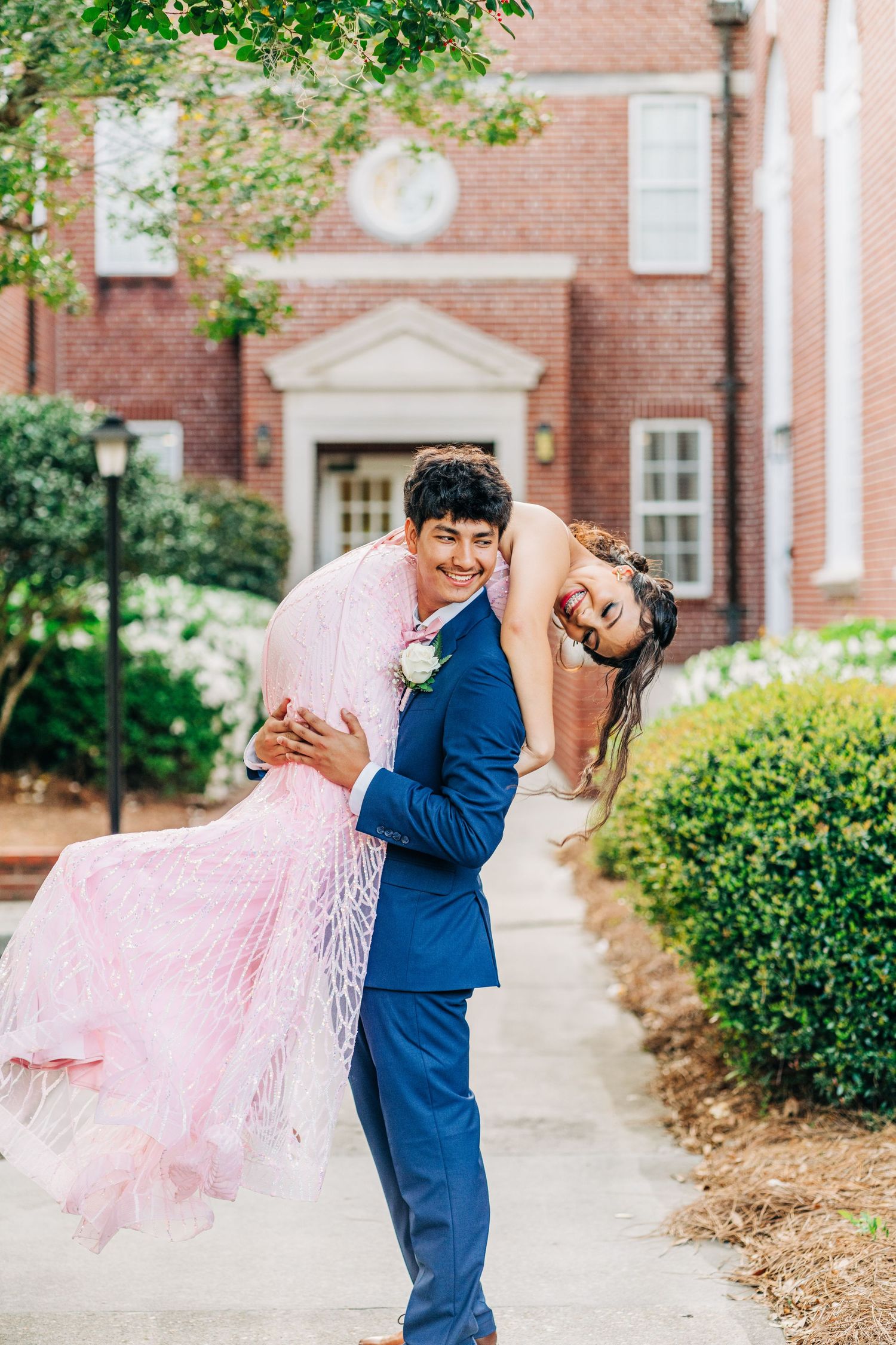 A couple in wedding attire embrace playfully on a brick walkway.