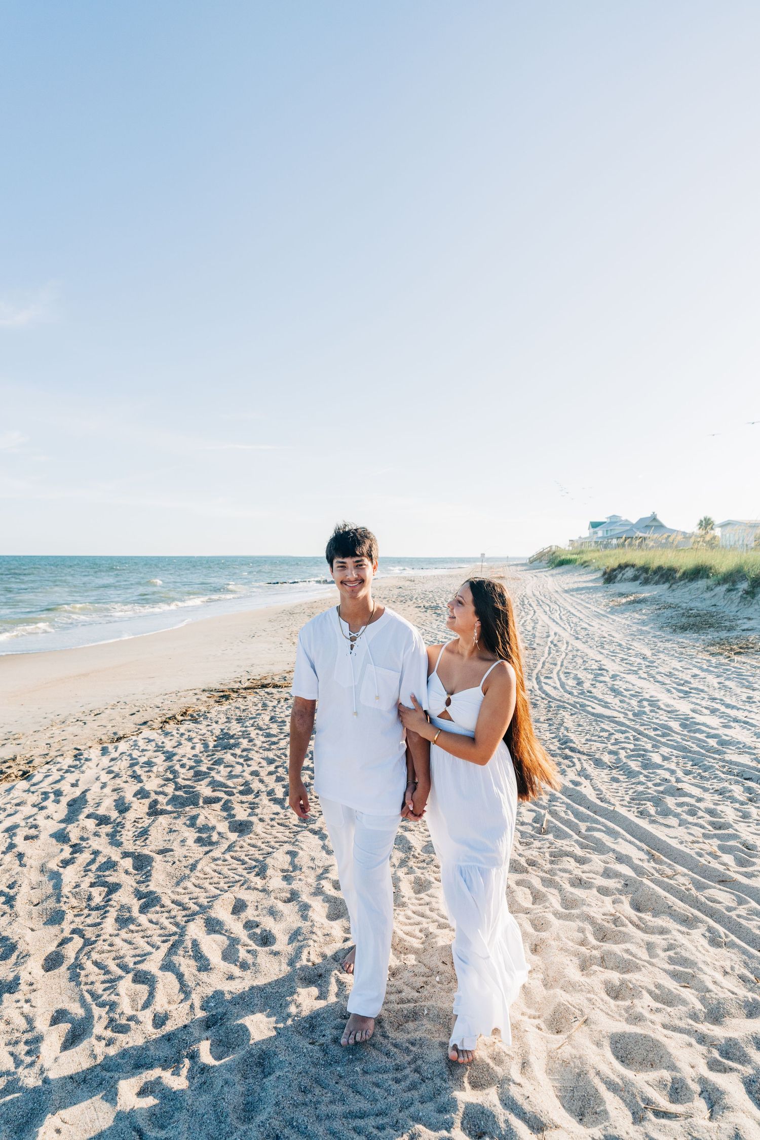 A couple in white outfits walks together along a sunny beach with coastal homes visible in the background.