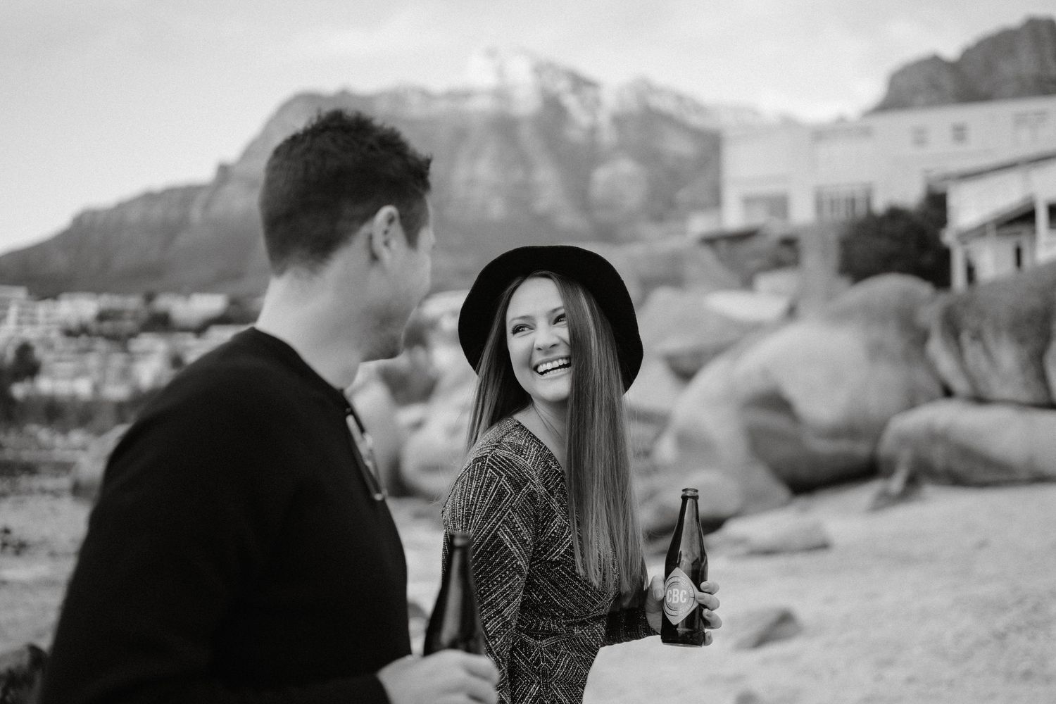 Two people sharing drinks on a beach with rocky cliffs and mountains in the background, captured in black and white.
