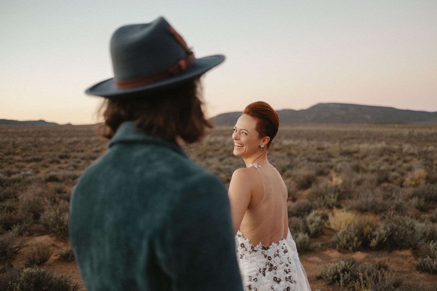 Desert engagement photography session with couple in boho dress and cowboy hat posing at sunset against desert landscape.