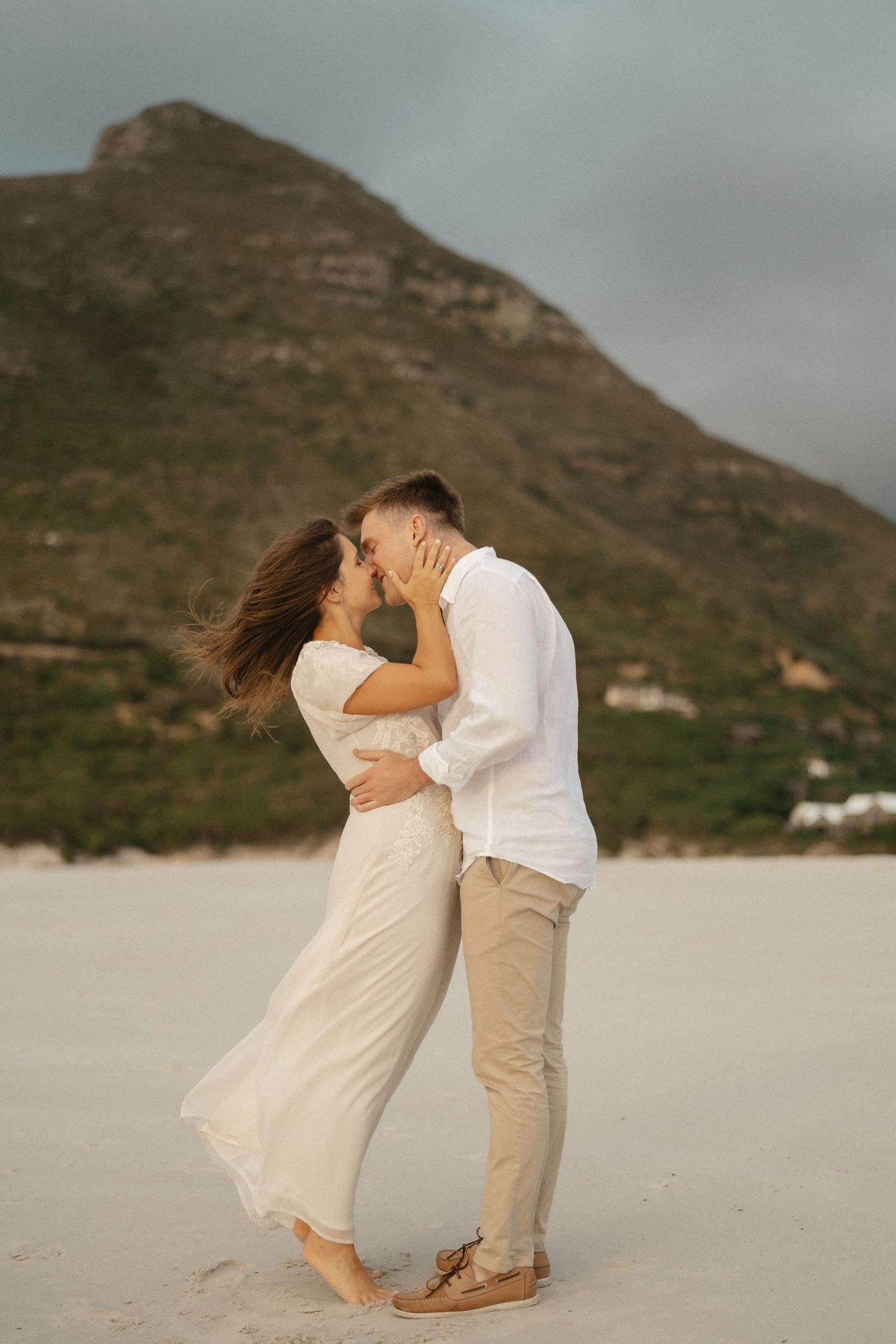 A romantic couple embraces on a beach with a dramatic mountain peak in the background at sunset.