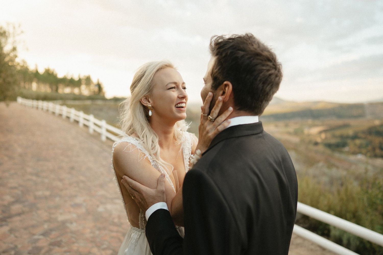A couple shares a joyful moment together on a dirt path with rolling hills and white fence in the background at sunset.