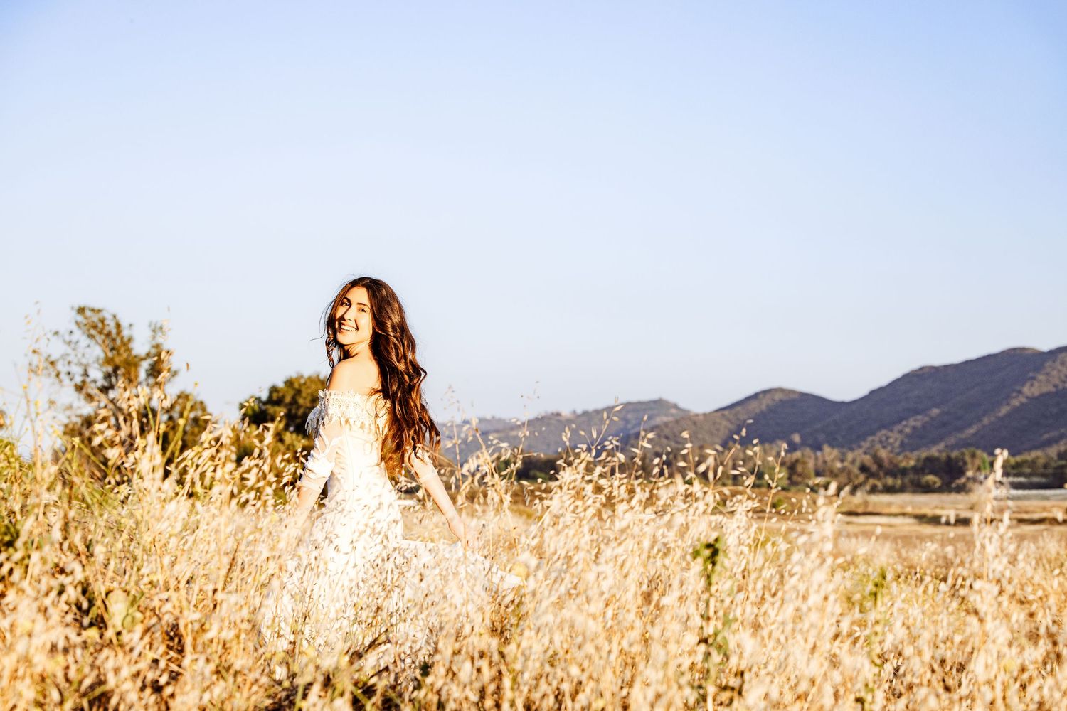 A person in a white dress poses gracefully in a golden field with mountains in the background on a sunny day.