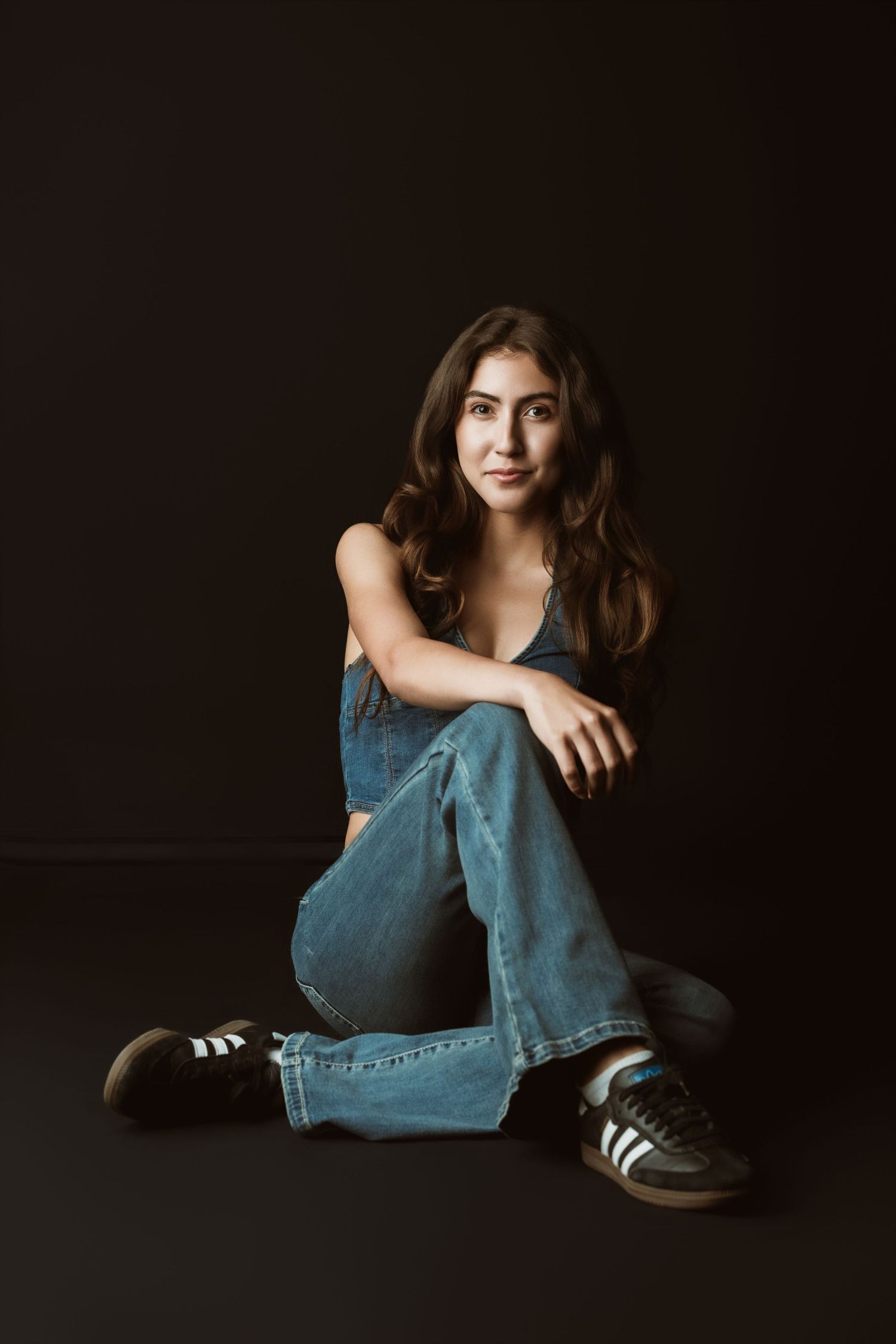 Dramatic studio portrait of a person in denim overalls and sneakers sitting cross-legged against dark background.