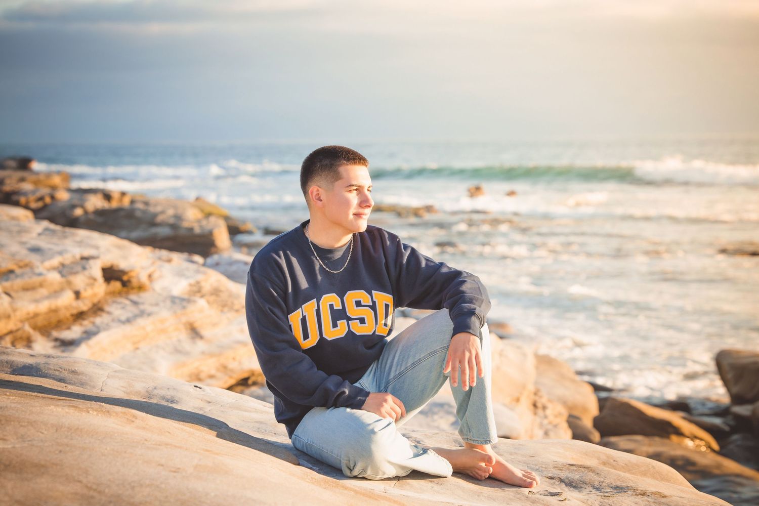 Person wearing a UCSB sweatshirt sits relaxed on coastal rocks during golden hour with ocean waves in the background.