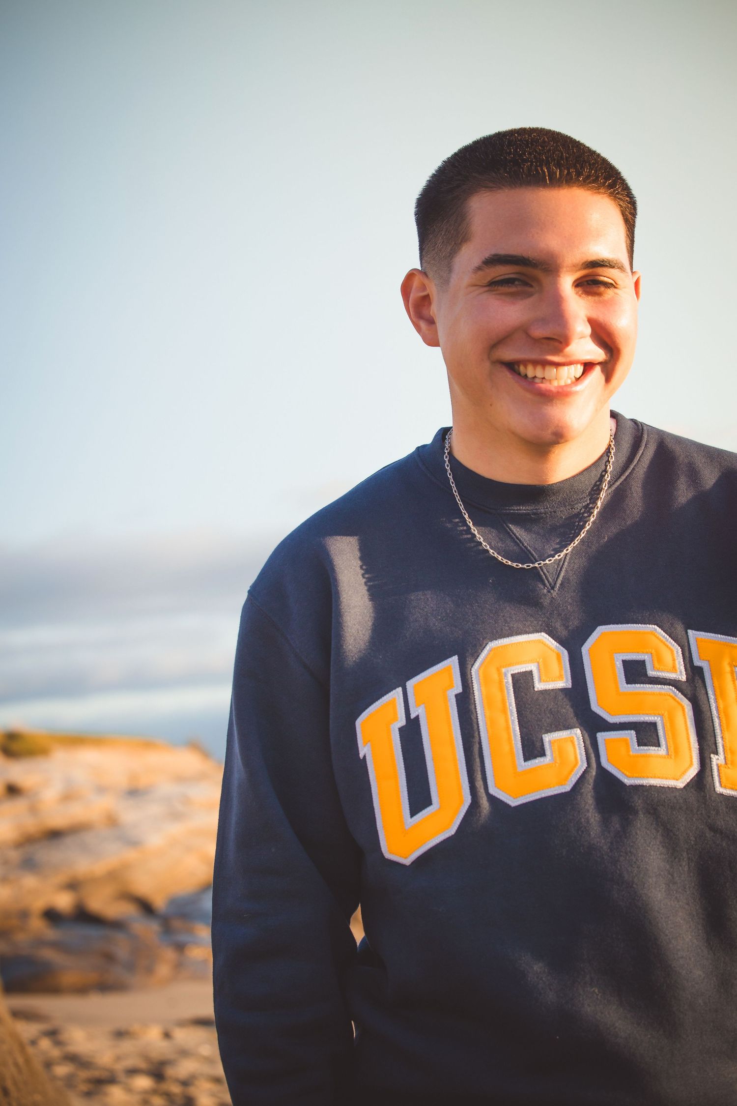 Someone in a dark blue UCSB sweatshirt smiles while standing by a rocky coastal area on a bright day.