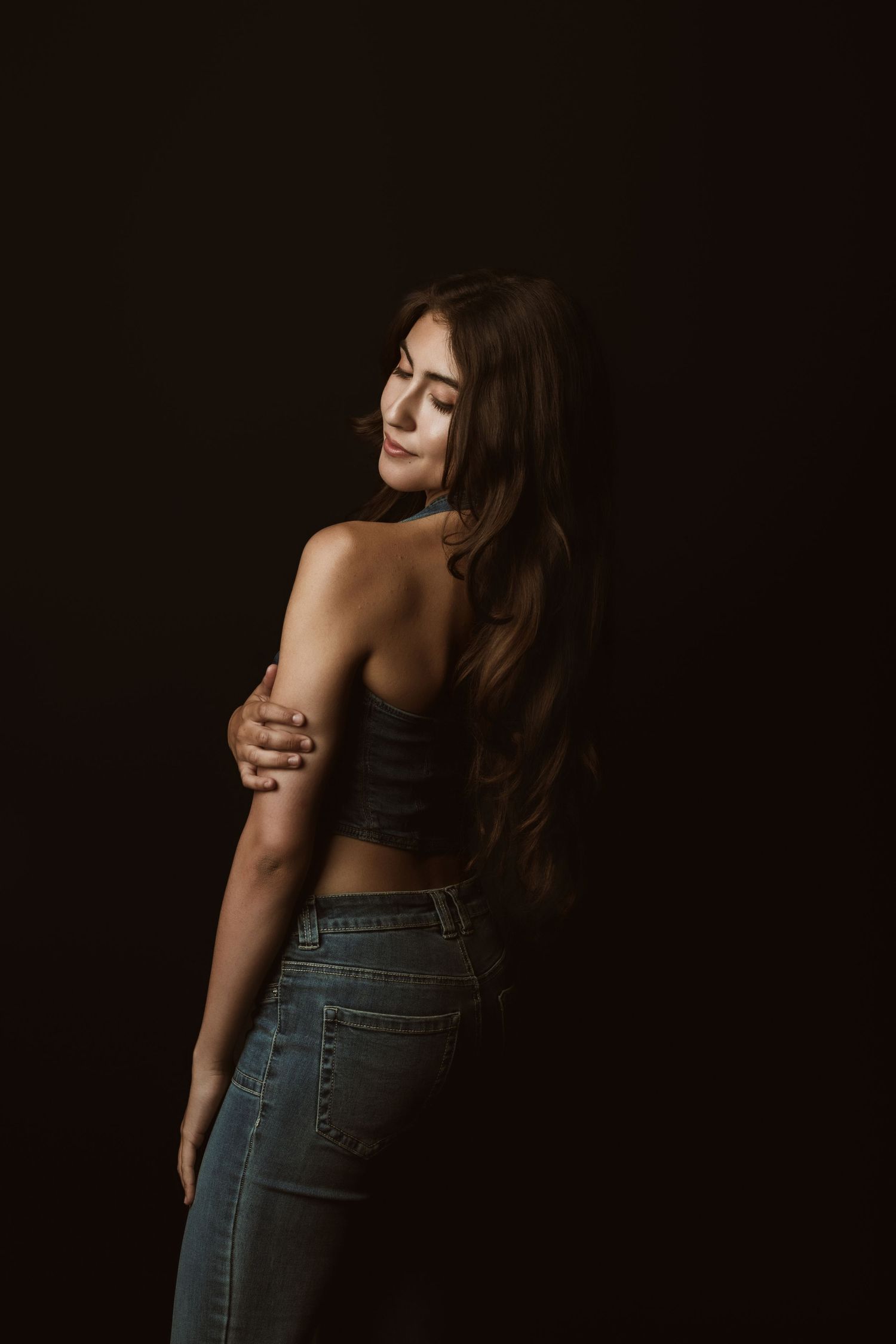 Moody portrait shot showing a person in jeans and crop top posing against dark background with dramatic lighting.