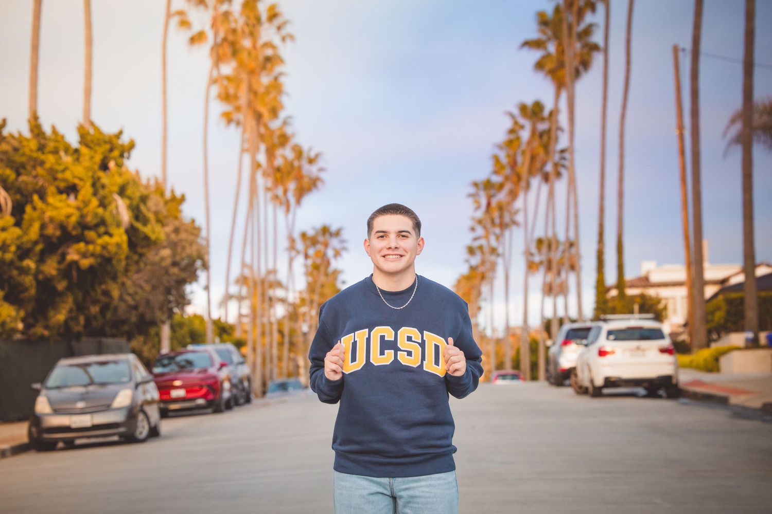 Person wearing a navy UCSD sweatshirt stands on palm tree-lined street at sunset with parked cars in background.