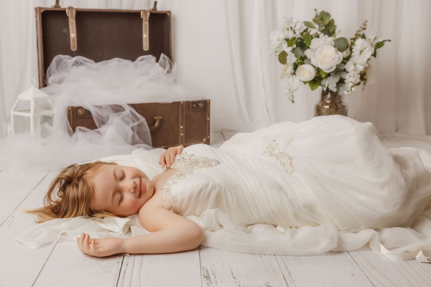 Dreamy bridal portrait with person lying on white floor next to vintage suitcase and floral arrangement.