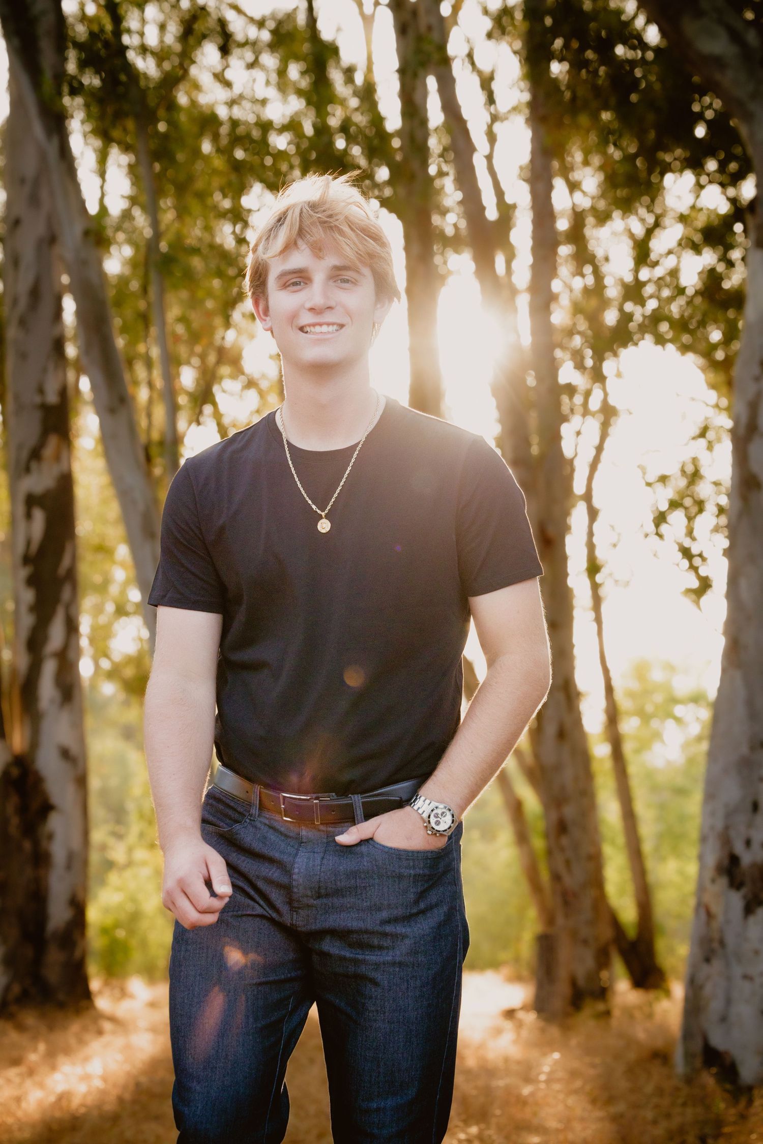 Portrait of someone in a black t-shirt and jeans standing outdoors in a sunlit wooded area at golden hour.