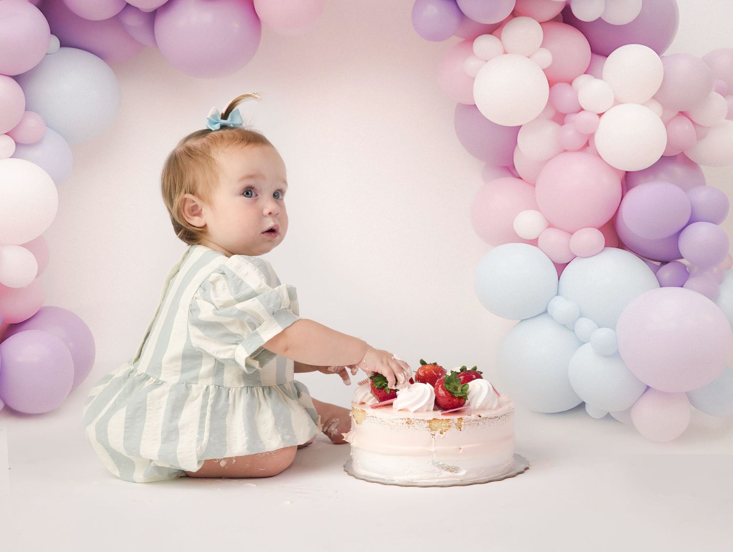 Baby in white dress celebrates with cake surrounded by pastel balloon arch backdrop.