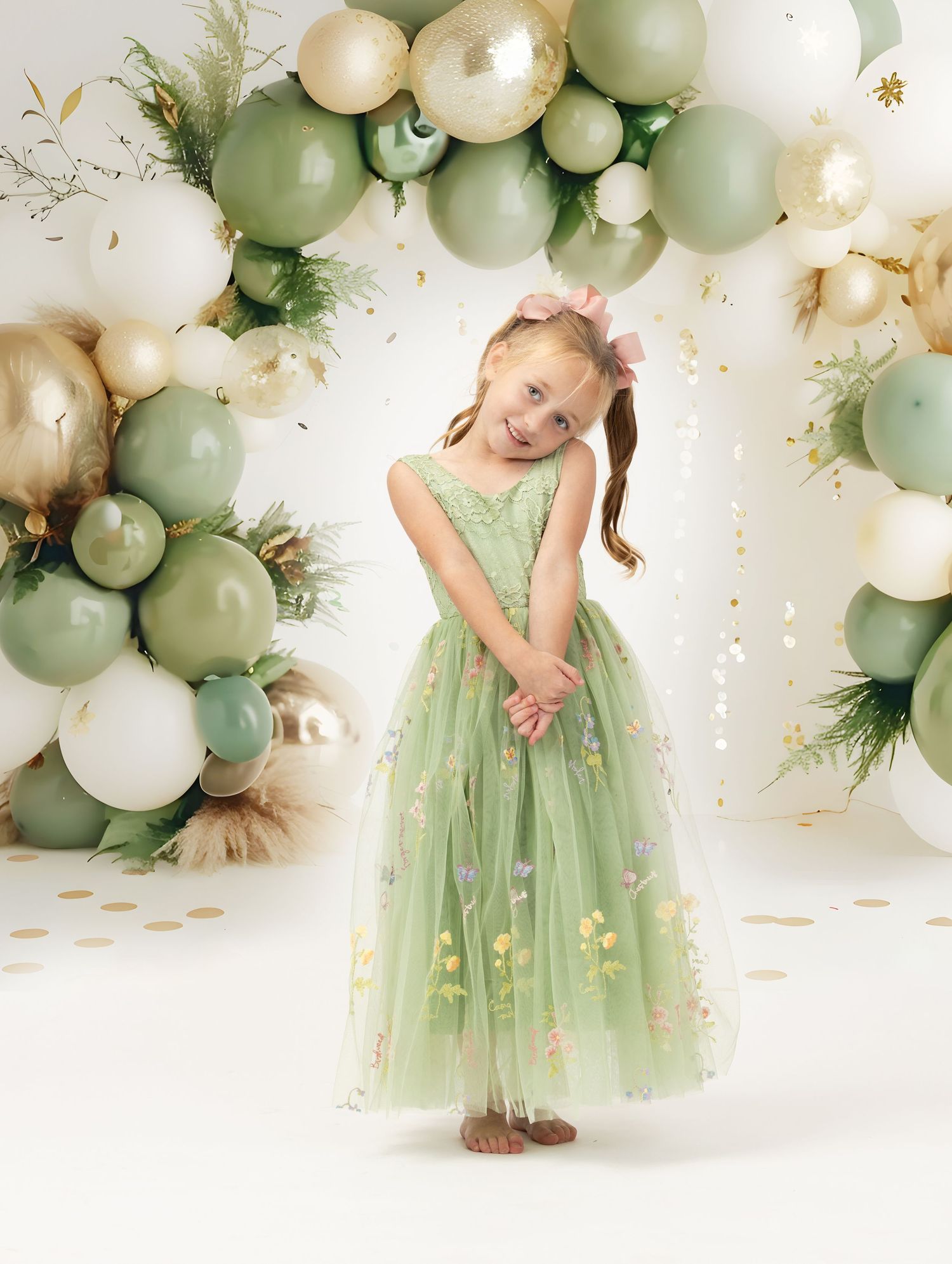 Sage green and gold balloon arch with eucalyptus accents creates an elegant party backdrop against a white wall.