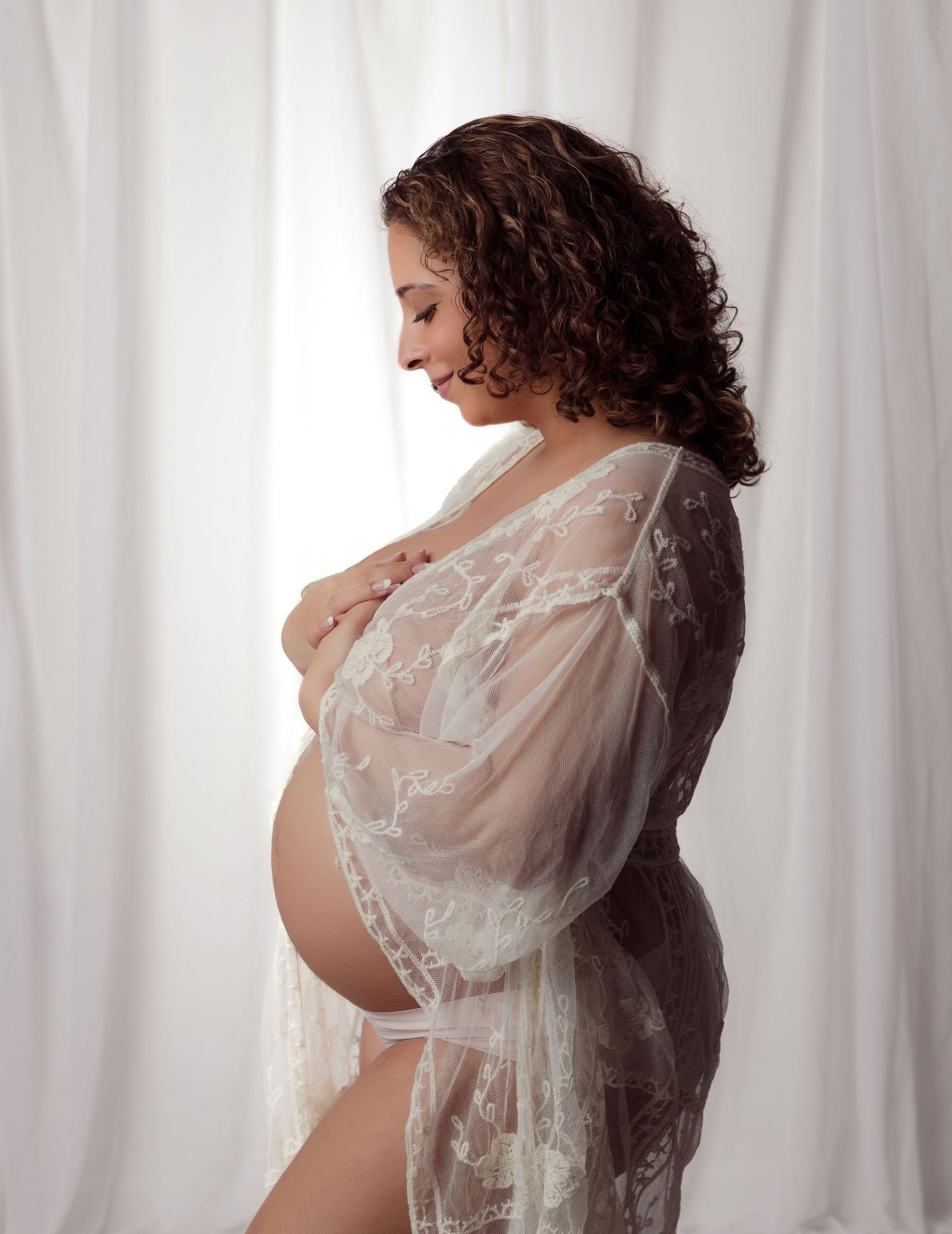 A pregnant woman in white lace lingerie and sheer robe poses in profile against a light curtained backdrop.