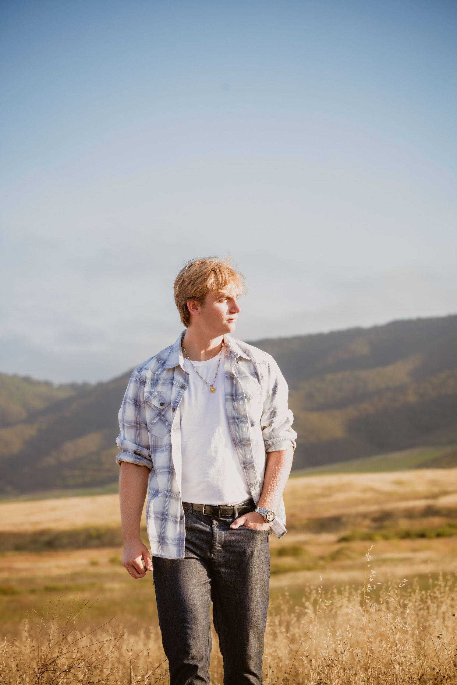Person wearing plaid shirt and jeans stands in golden grassland with rolling hills in background on sunny day.