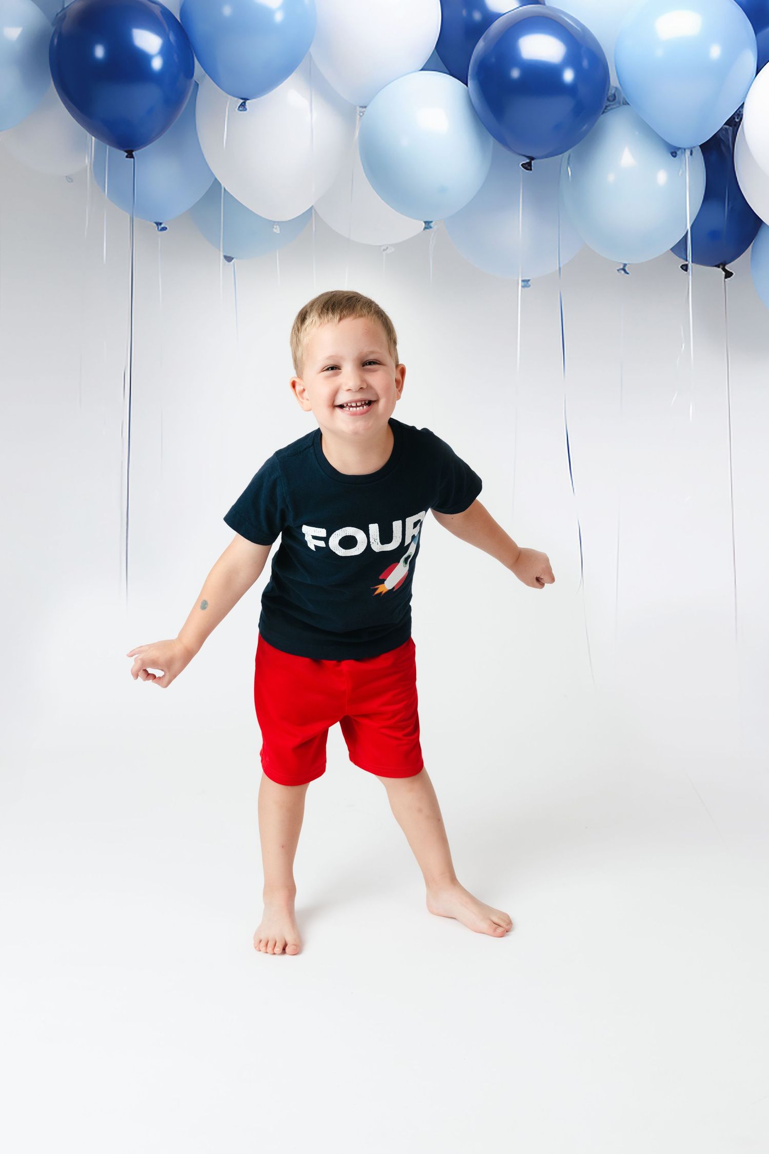 Toddler in black shirt and red shorts stands playfully against white background decorated with blue balloons.