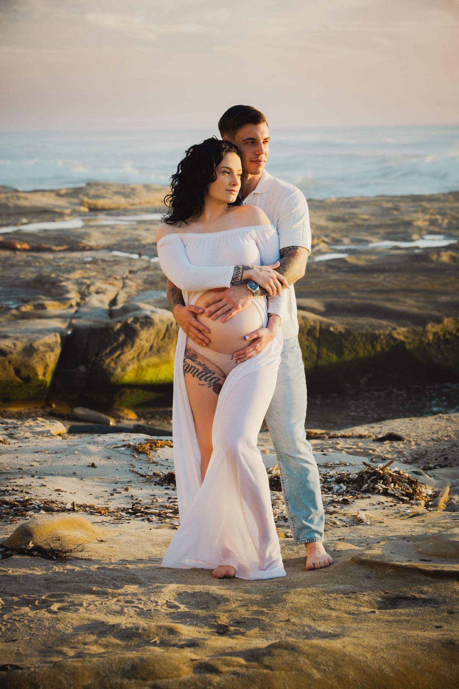 A romantic couple embraces on rocky coastal terrain during golden hour sunset lighting.