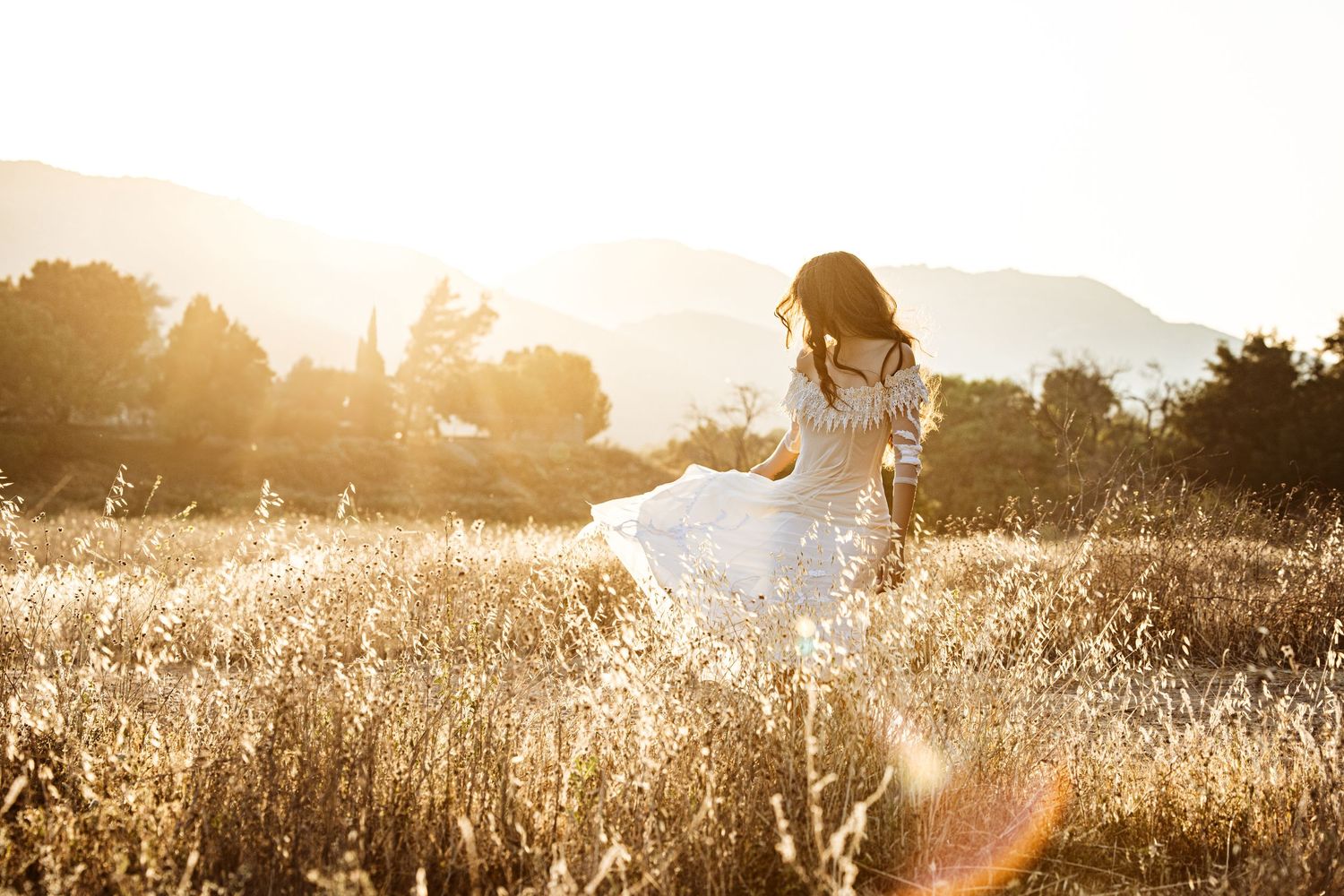 A woman in white dress walks through a golden sunlit field during sunset.