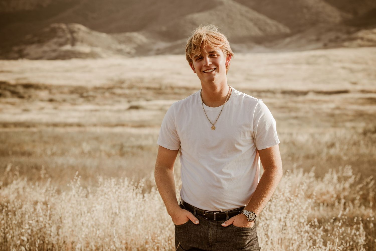 Person in a white t-shirt and jeans stands smiling in a golden field with mountains in the background.