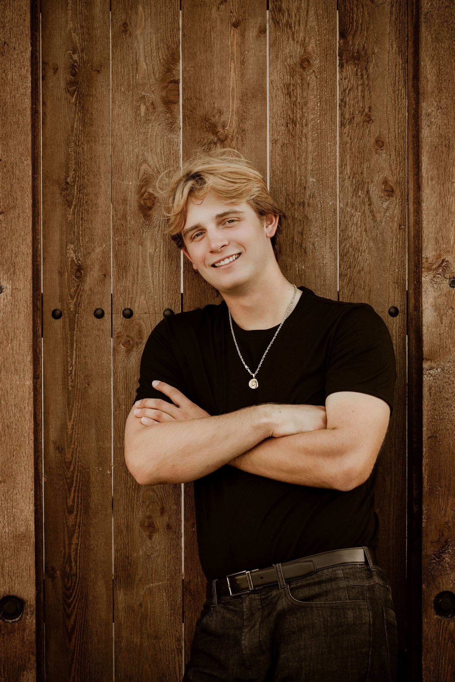 Casual portrait of someone in a black t-shirt and necklace leaning against rustic wooden door background.