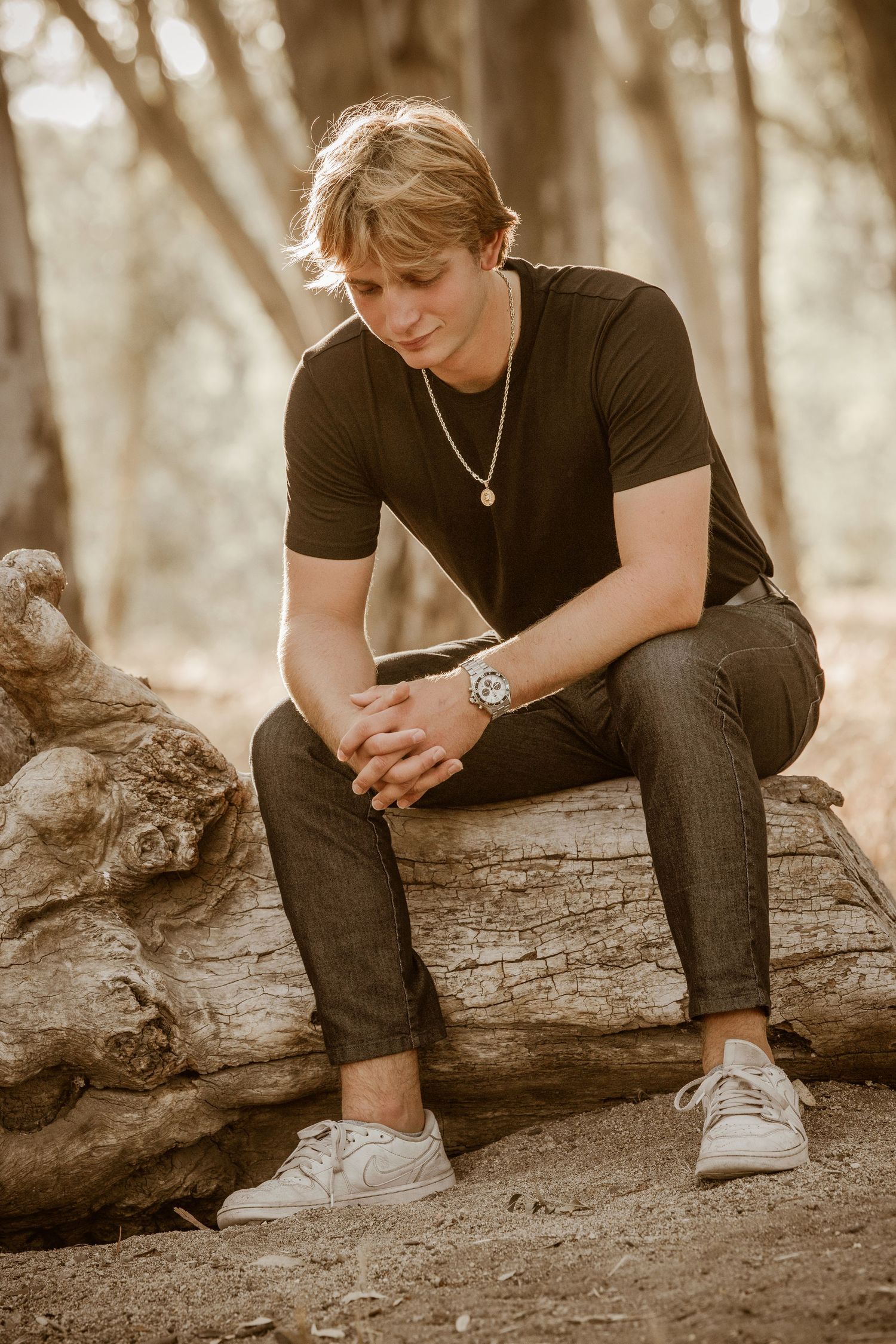 A person in a black t-shirt and jeans sits thoughtfully on a fallen log in a sunlit forest setting.