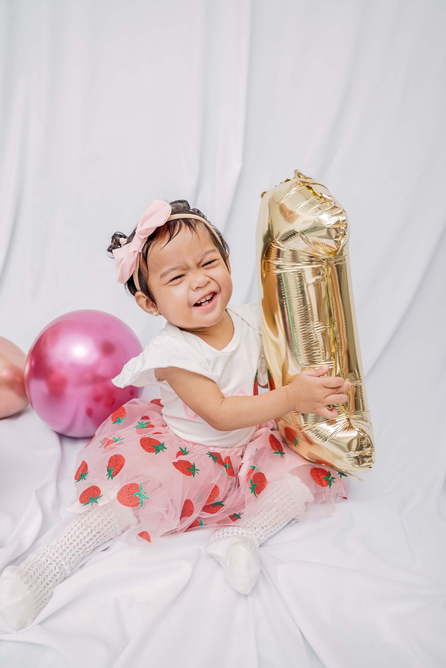 Laughing child in white dress lies on floor with gold balloon and pink decorations during first birthday photoshoot.