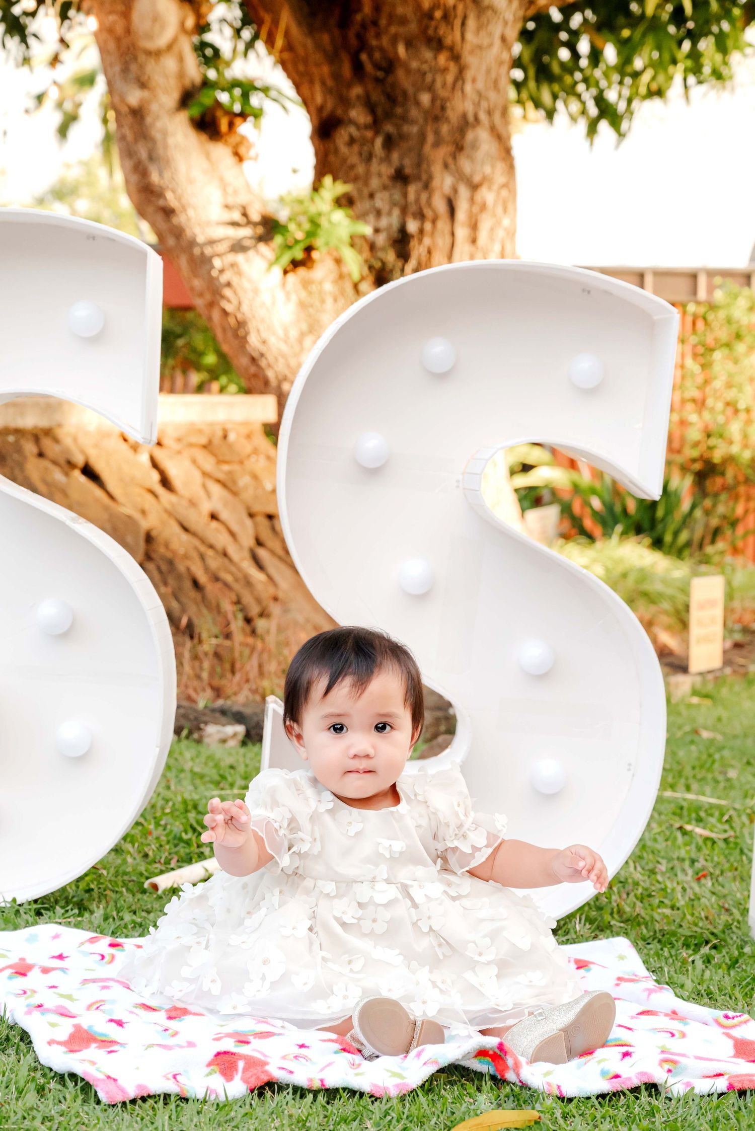 A small child in a white lacy dress sits on a floral blanket near large decorative letters in an outdoor garden setting.