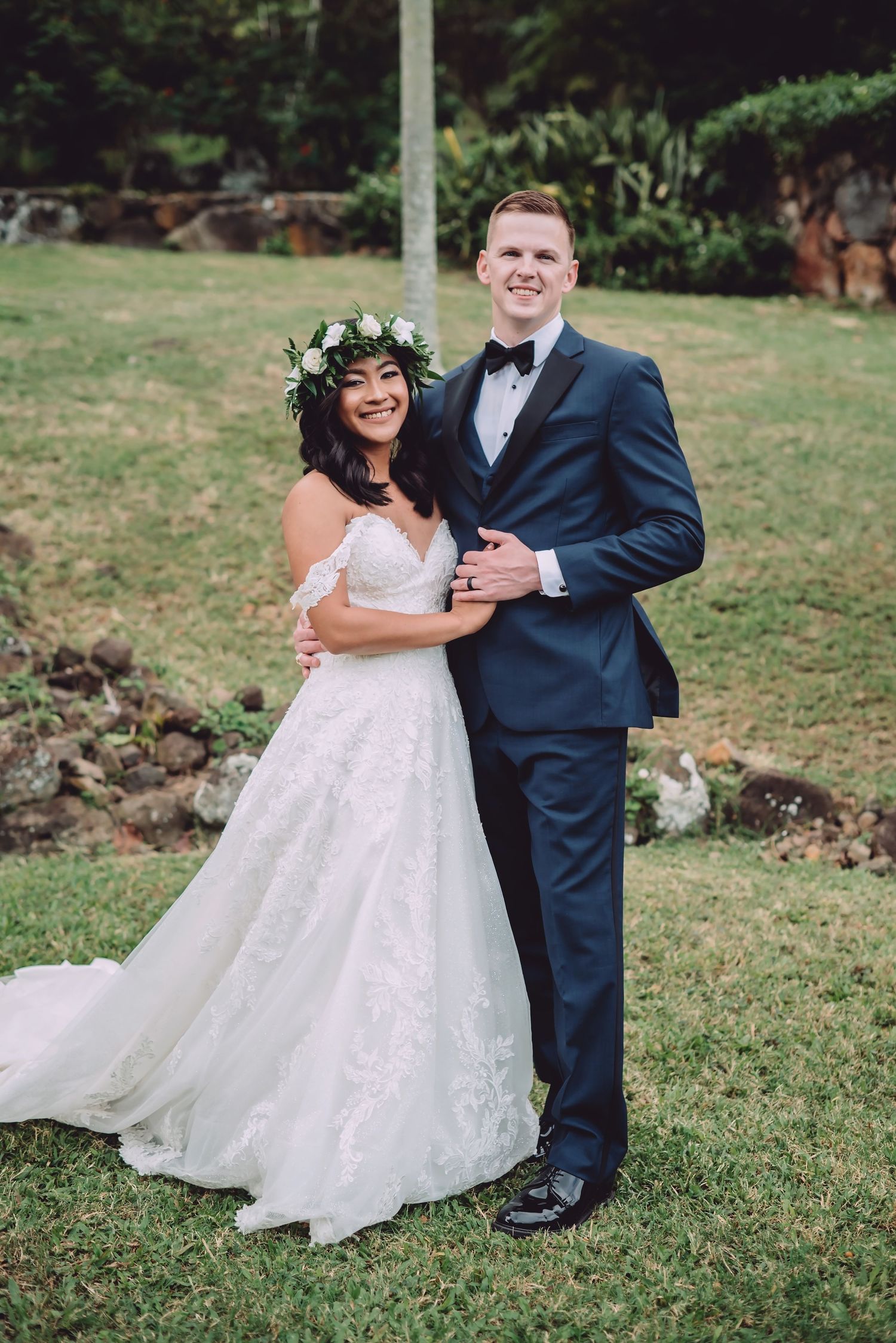 A wedding couple poses outdoors on a lawn, the bride wearing a floral crown and flowy white dress, groom in navy suit.