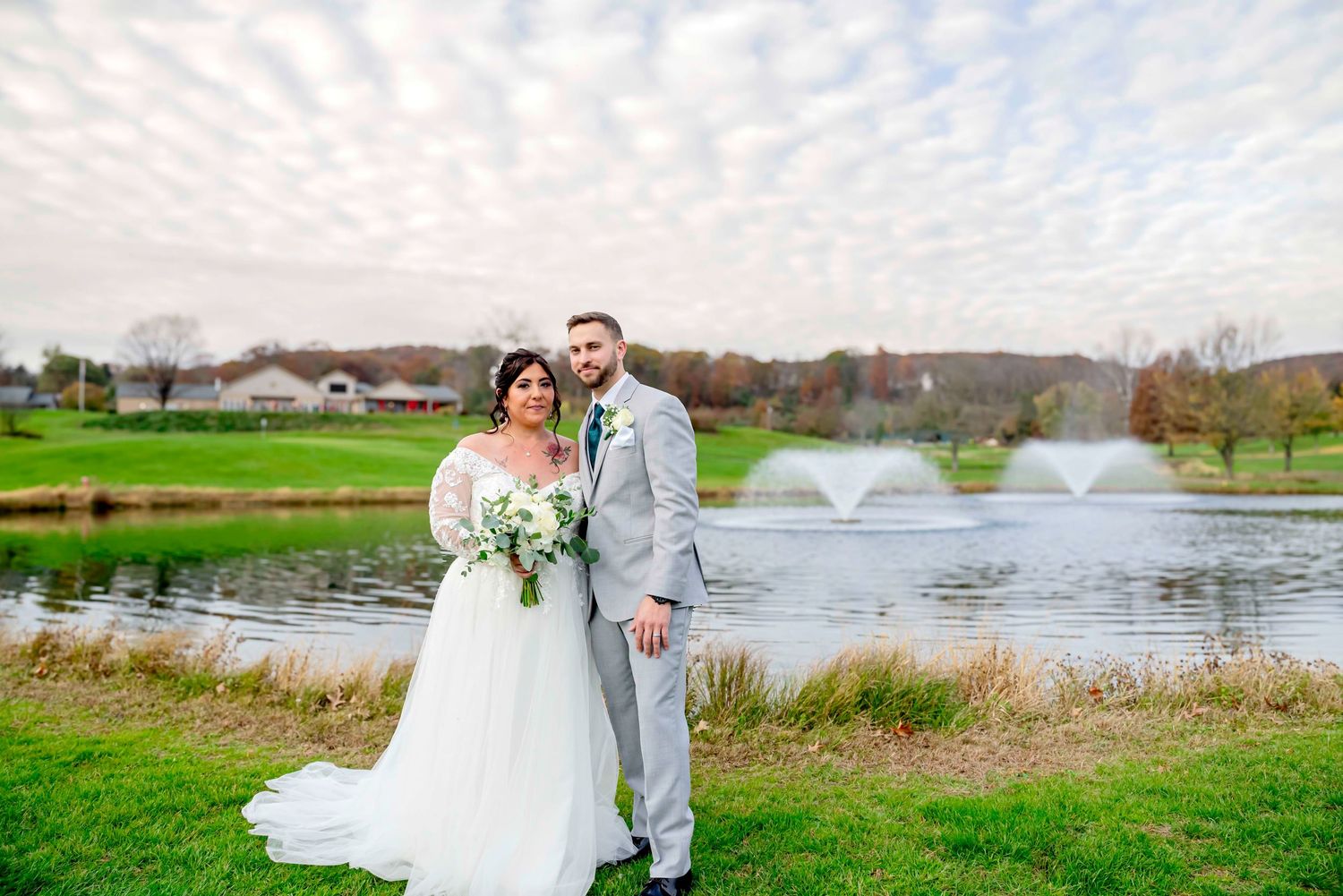 A newlywed couple poses together by a scenic pond with fountains at a golf course on their wedding day.