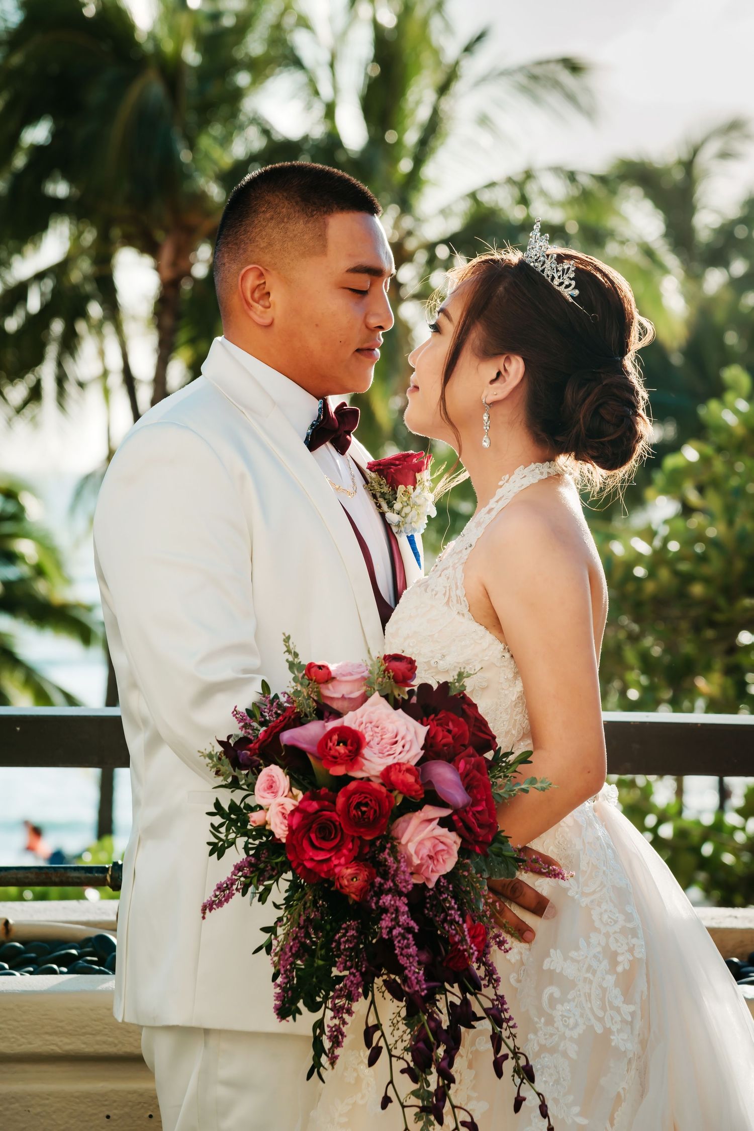 A newlywed couple shares an intimate moment on a tropical terrace with a stunning floral bouquet of pink and red roses.
