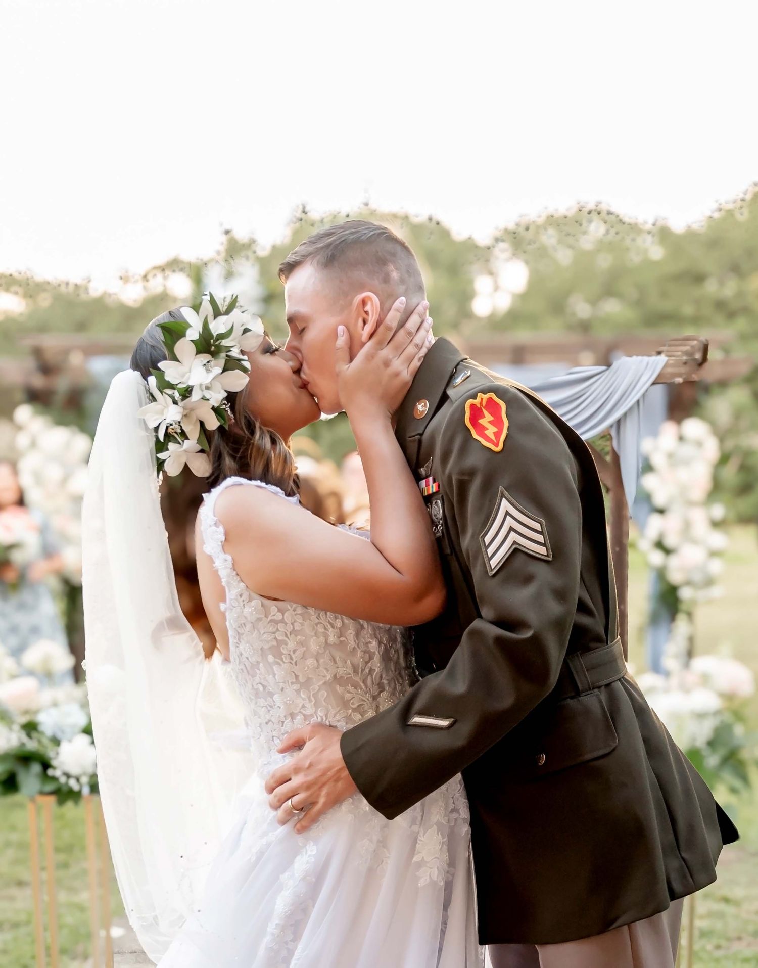 A couple shares romantic kisses during an outdoor wedding ceremony surrounded by summer greenery.