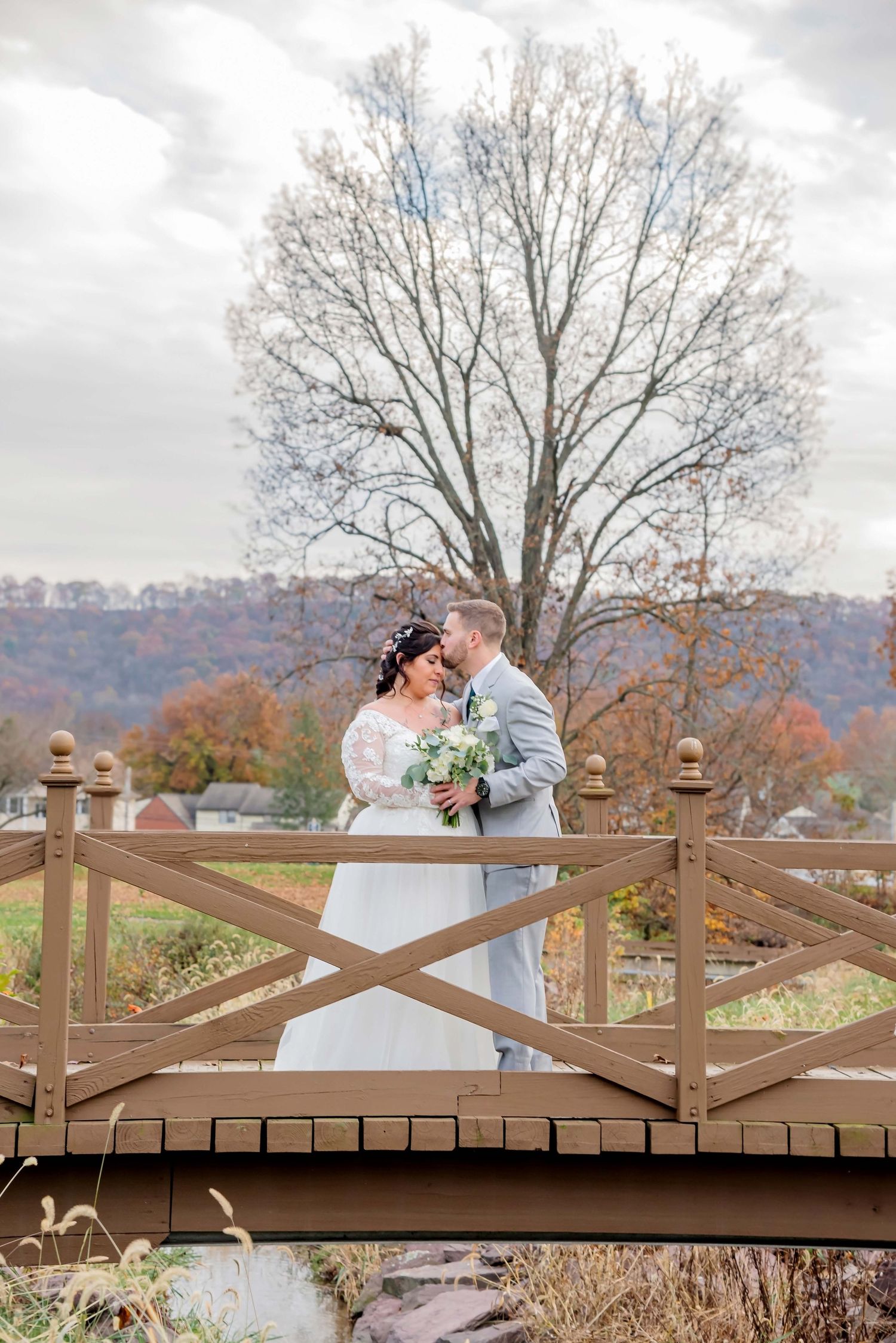 Couple embracing on wooden bridge with autumn foliage and mountains in background.