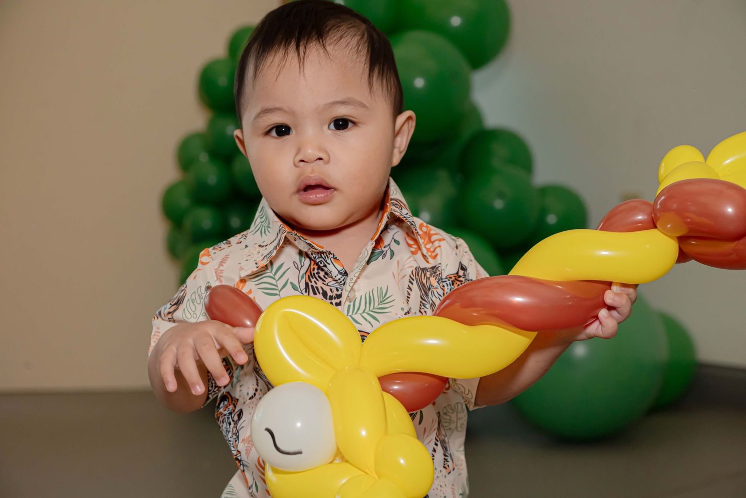 Child holding a yellow balloon animal toy at a party with green balloons in the background.