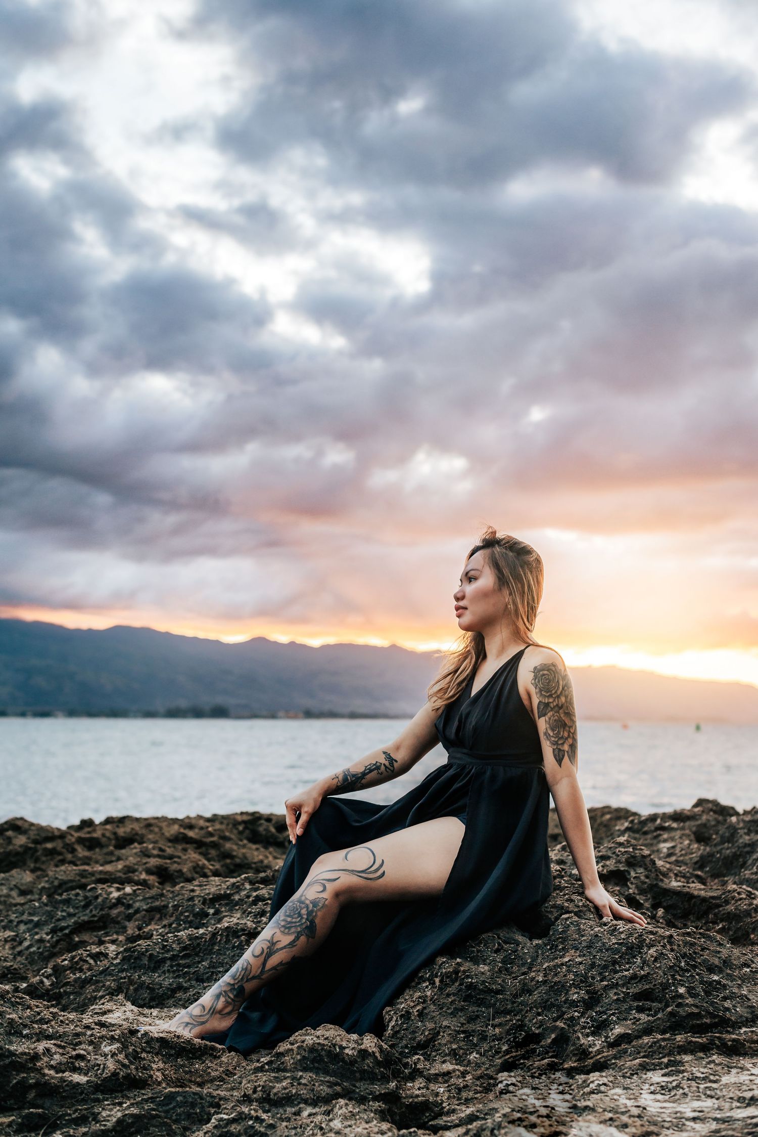 A person in a black dress with tattoos sits on coastal rocks at sunset with dramatic cloudy skies and mountains in the distance.