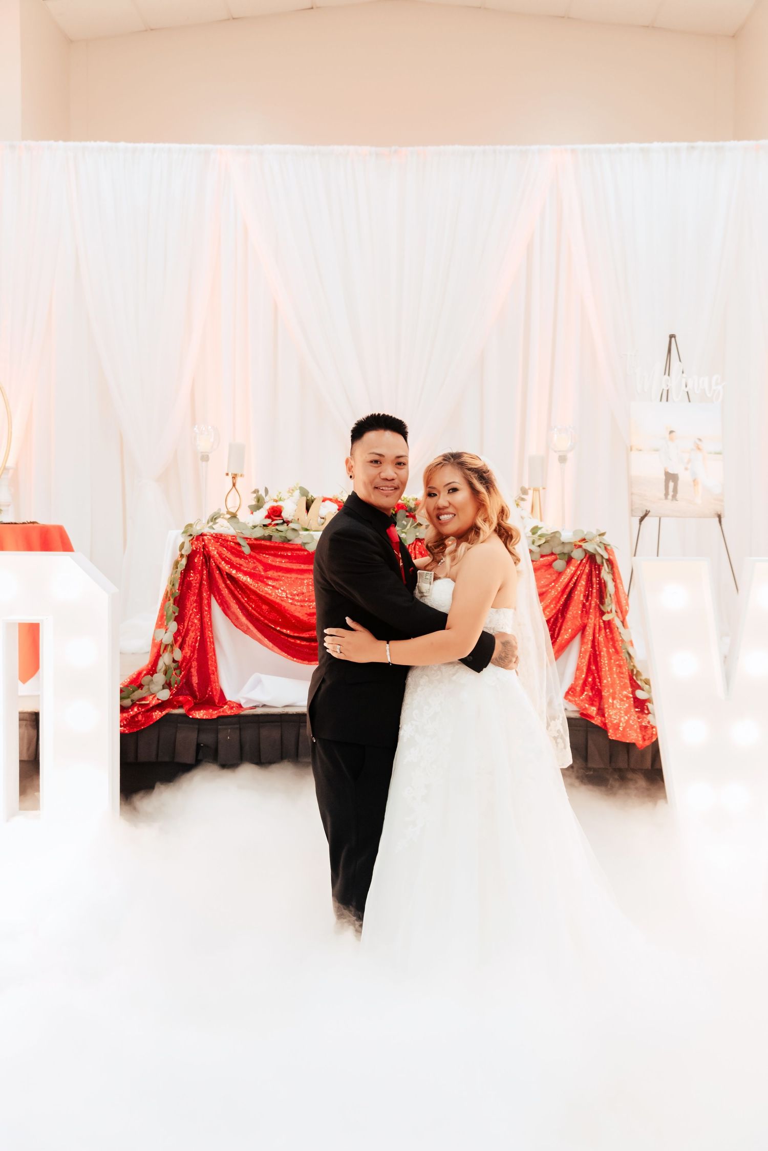 Newlyweds embrace in front of elegant reception decor with white draping and red sequin tablecloth.