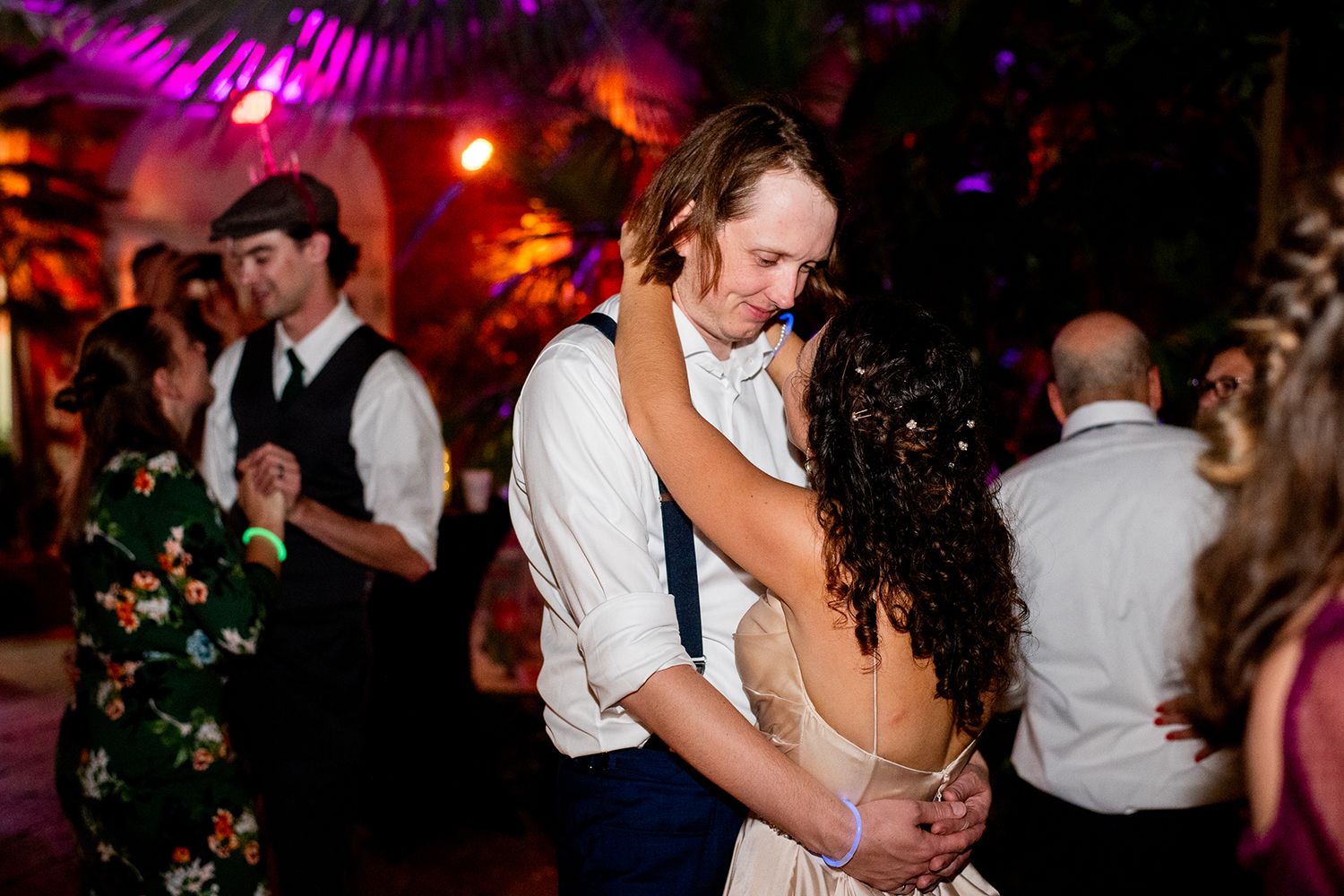 Wedding guests dance and celebrate together on a dimly lit dance floor with red and purple lighting.