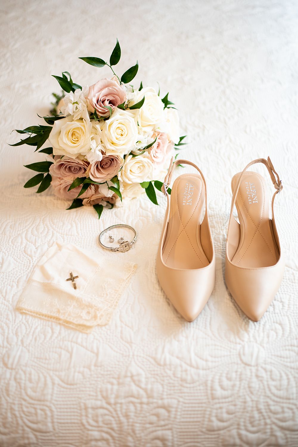 Elegant bridal shoes, pink and white roses, and wedding rings arranged on white textured fabric.