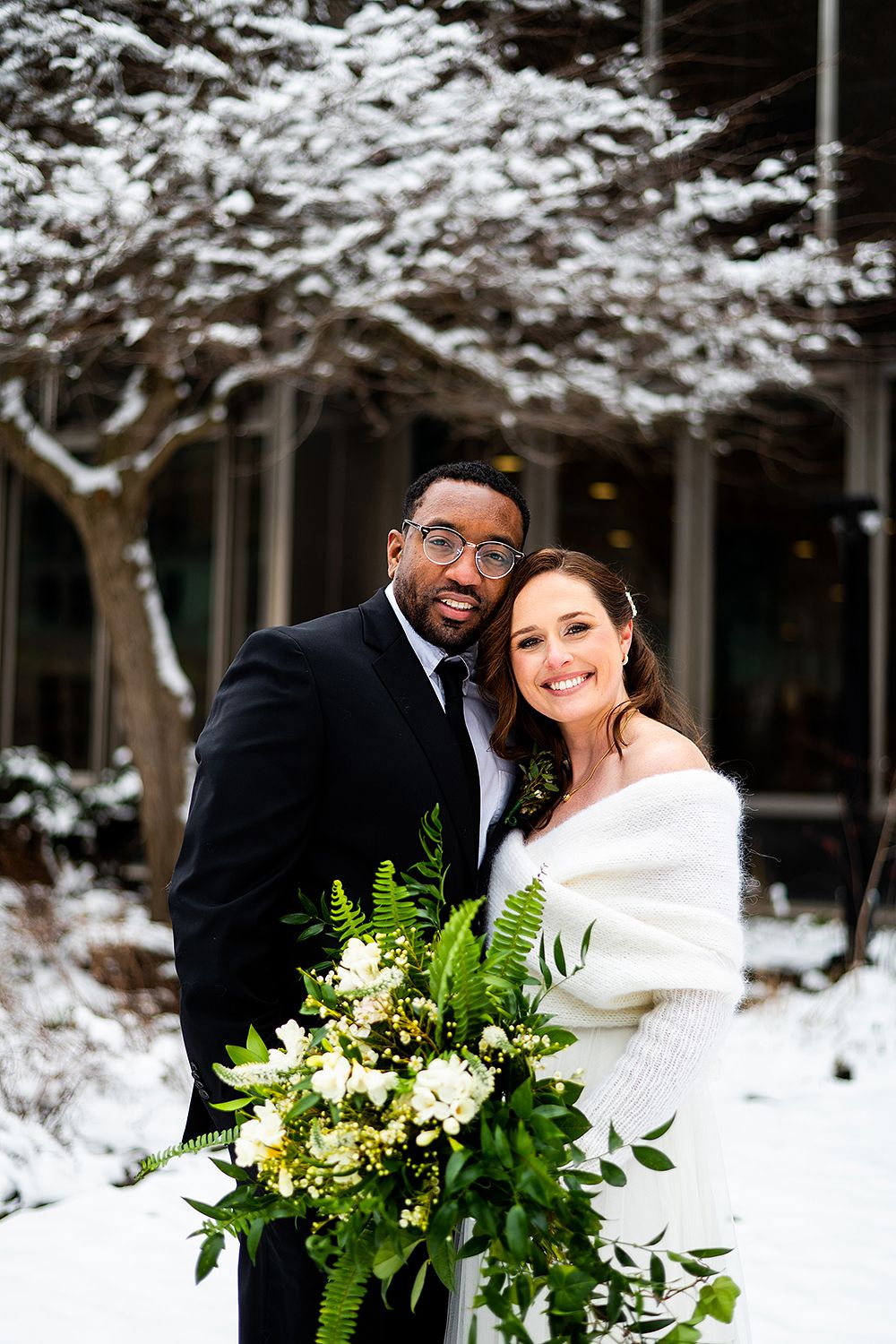 Happy couple embraces in snowy winter wedding scene with lush greenery bouquet of ferns and white flowers.