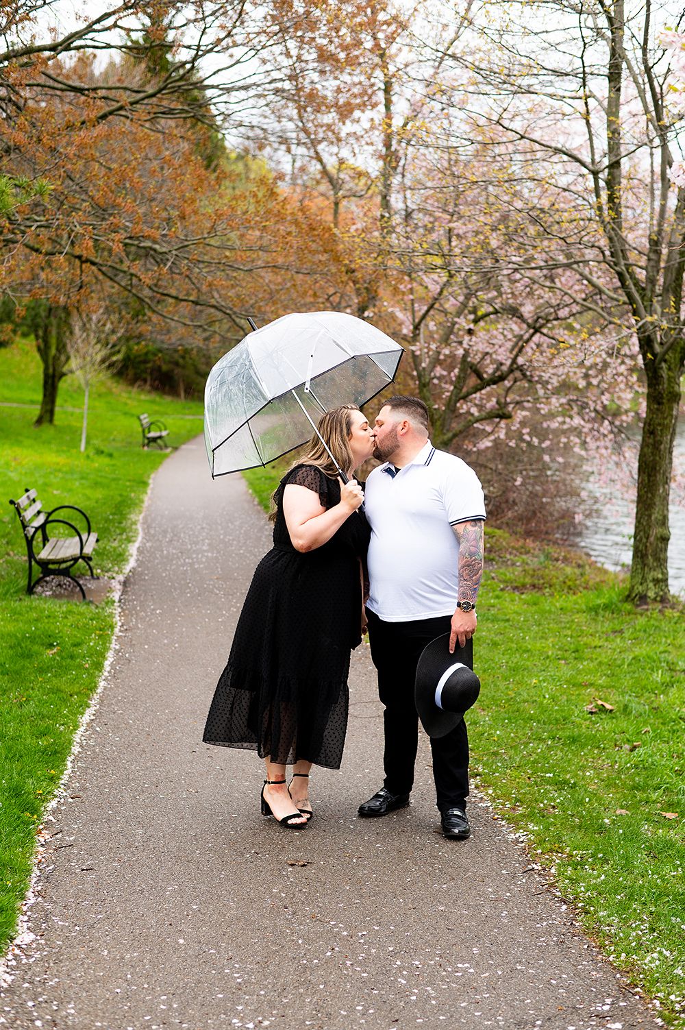 A couple shares a romantic moment under a clear umbrella on a park path surrounded by spring blossoms.
