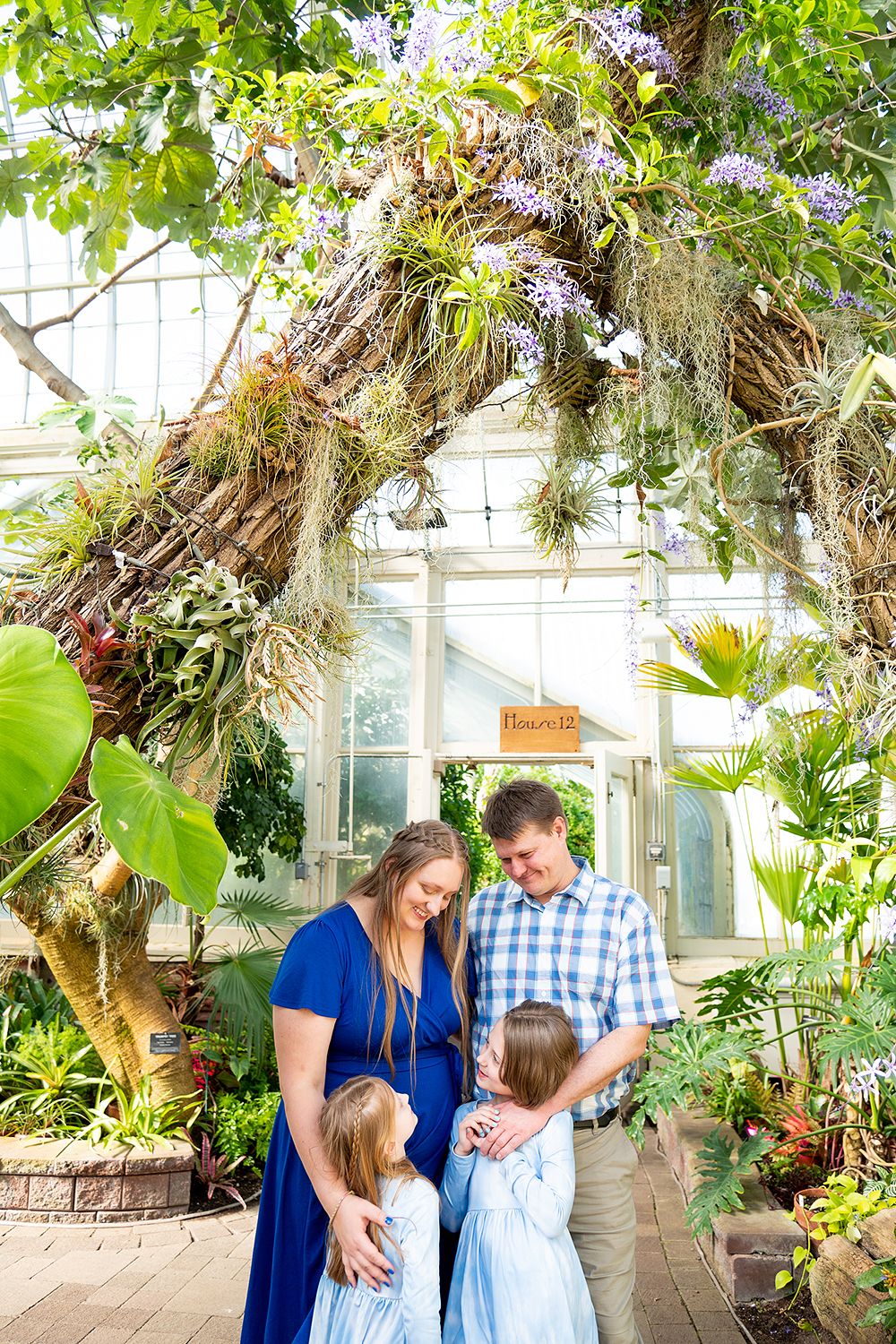 Family photographed together under a natural archway of tropical plants and flowers in a bright greenhouse setting.