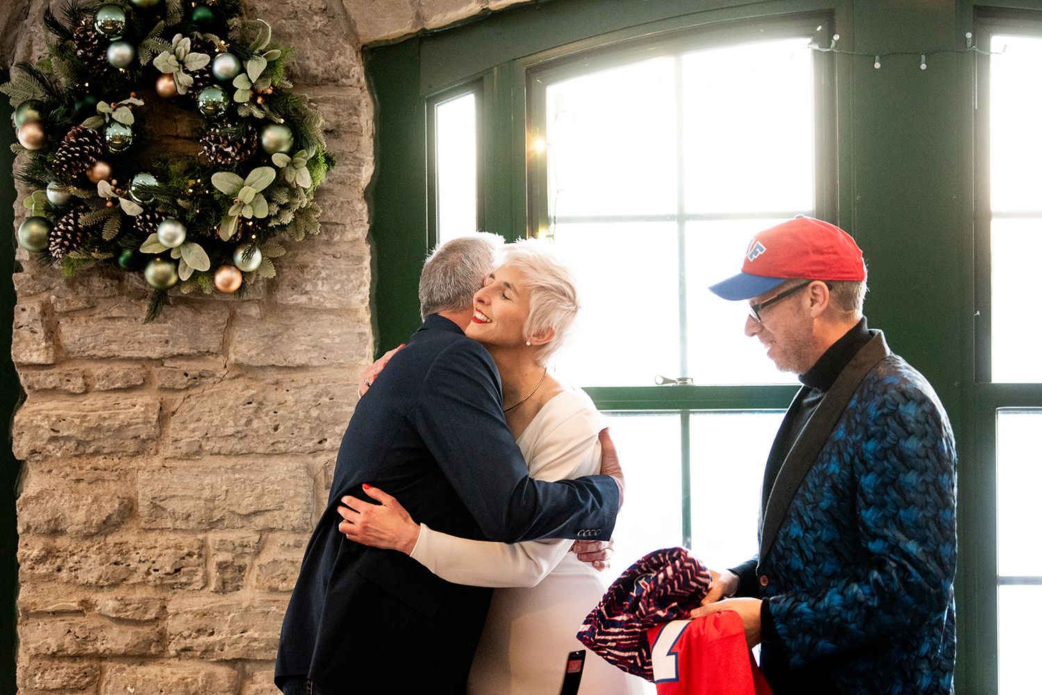 People gathered in a stone-walled room with natural light streaming through green-trimmed windows and festive decor.