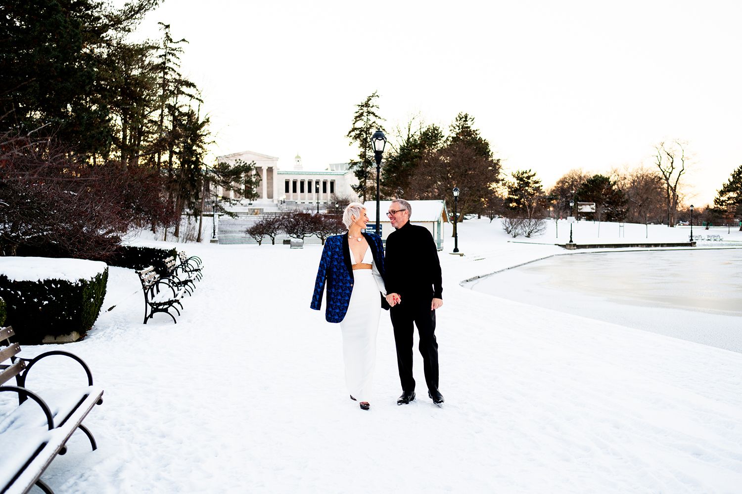 A couple walks together through a snowy landscape with a grand building visible in the distance.