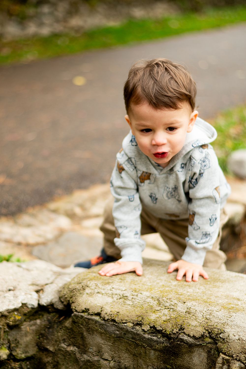 A young child in a gray hoodie explores a stone wall while playing outdoors on a paved path.